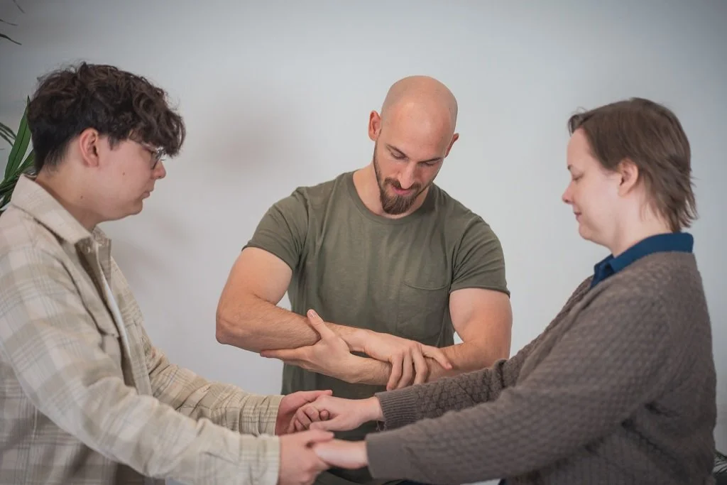 Three people holding hands in a circle with Adrian in a green t-shirt standing in the middle, the other two hold their hands in the background.