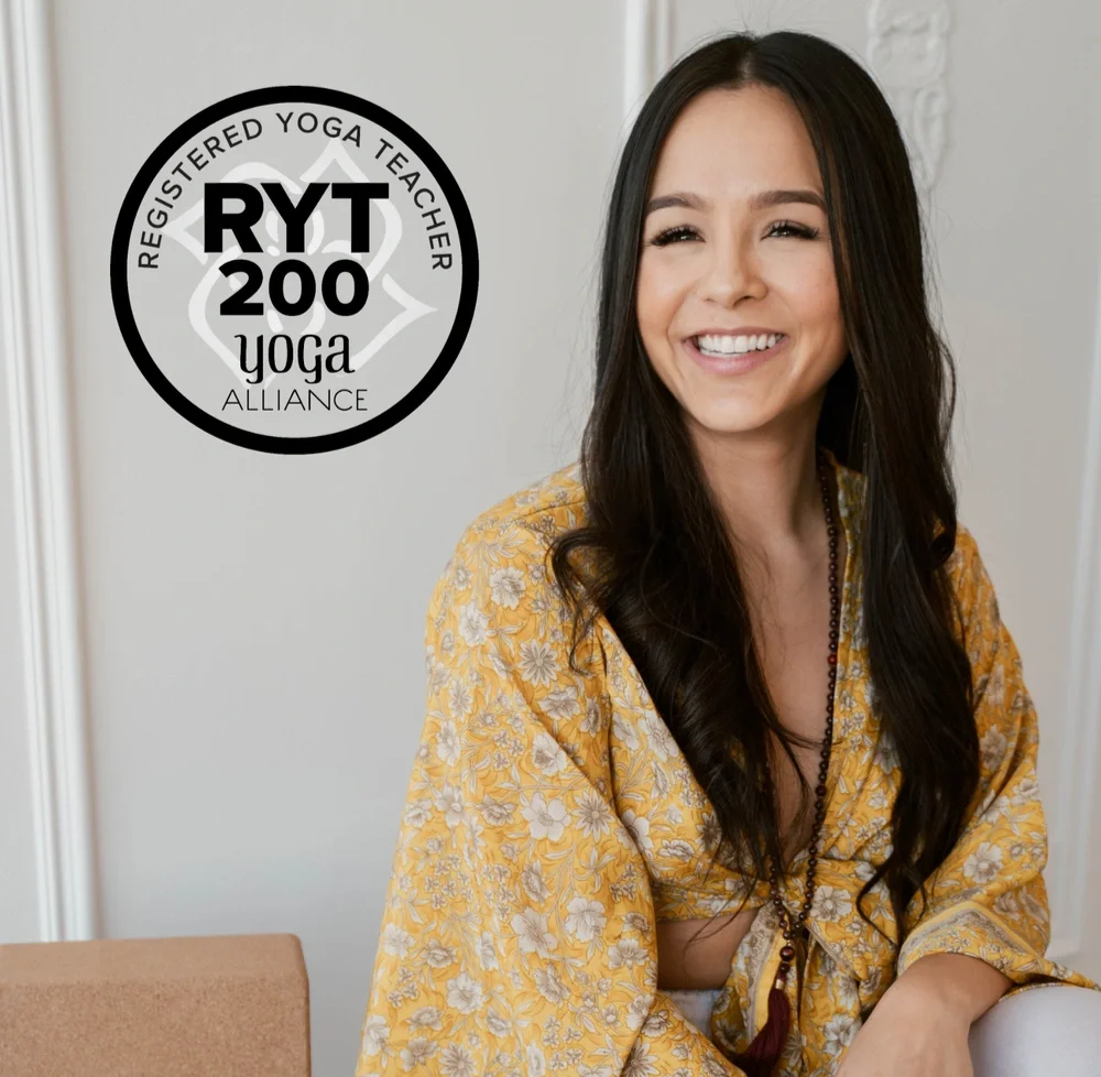 Smiling woman with long dark hair wearing a yellow floral shirt sitting indoors, with a yoga alliance RYT 200 badge on the wall beside her.