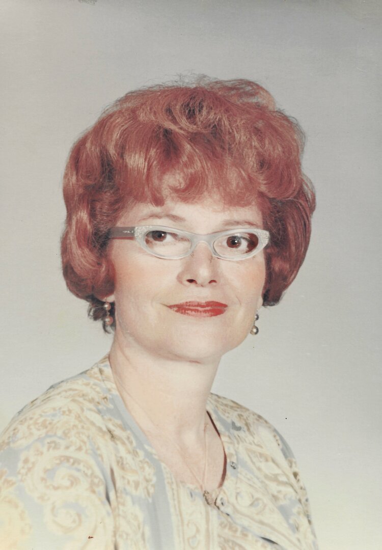 A woman with short, curly red hair, wearing glasses, earrings, and a patterned blouse, smiling slightly against a plain background.