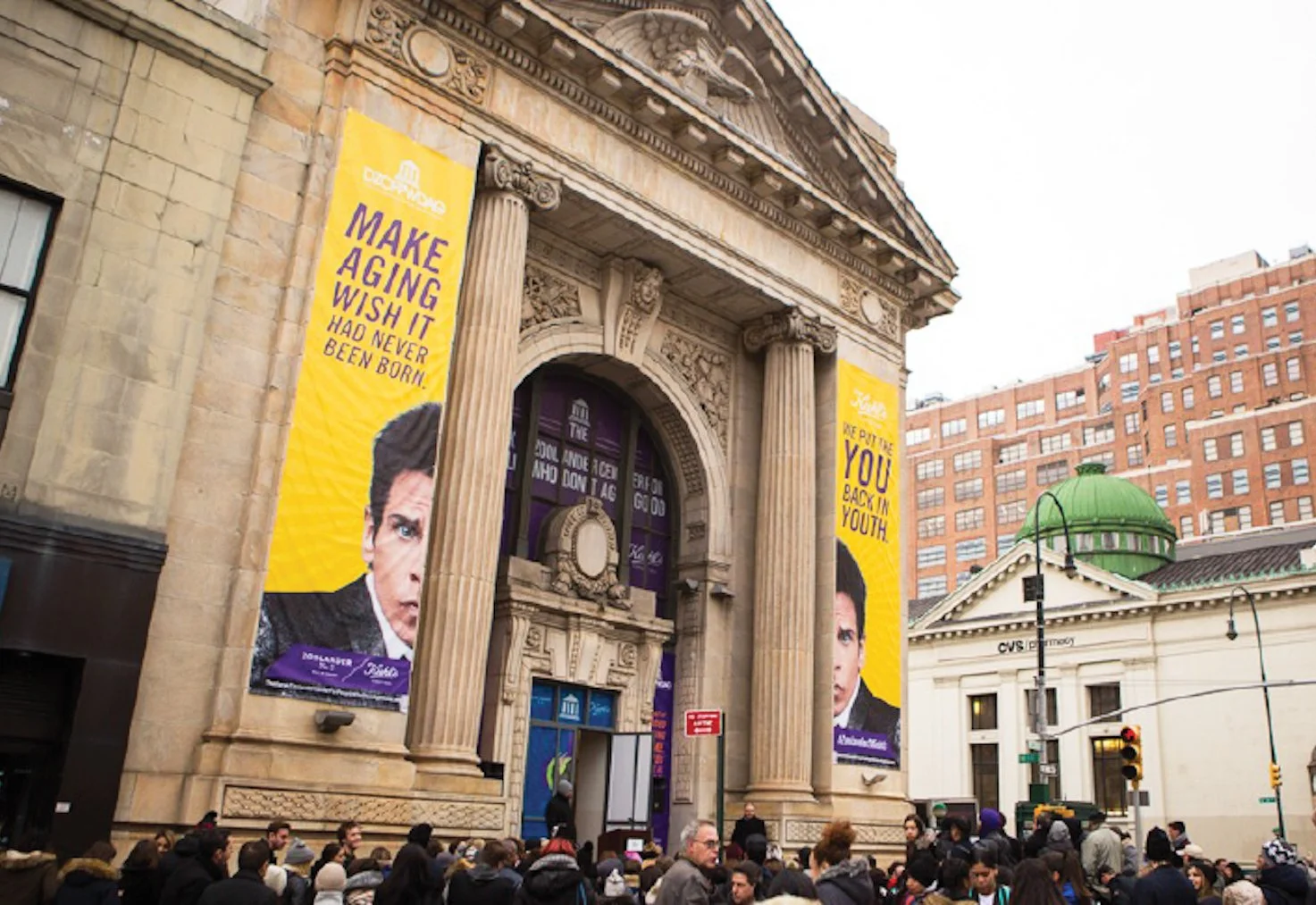 Crowd of people gathered outside a historic building with large columns and ornate architecture, featuring two tall yellow banners with a portrait of a man and the text 'Make aging wish it had never been born!'.