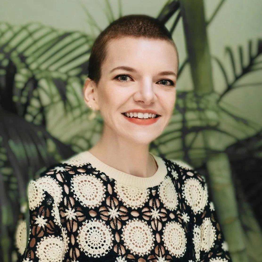 A woman with short hair smiling, wearing a crochet patterned black and cream sweater, standing in front of green tropical plants.