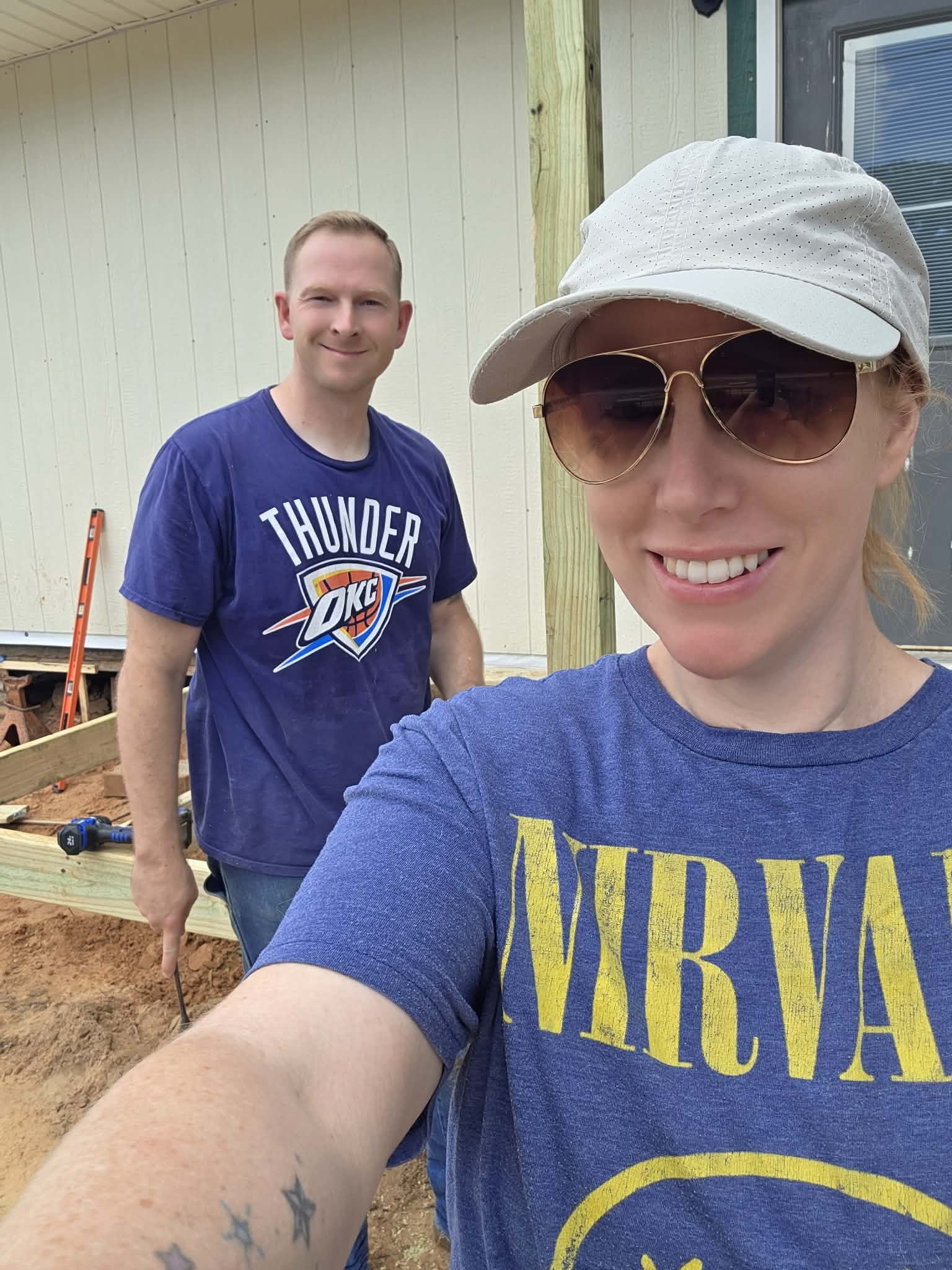 Two smiling women taking a selfie outdoors during daytime at a construction site, with wooden framing and tools in the background.