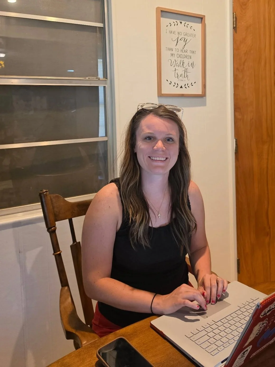 A woman with long brown hair sitting at a wooden table, smiling, working on a laptop, with a framed quote on the wall behind her that reads 'I have no greater joy than to hear that my children walk in truth.'