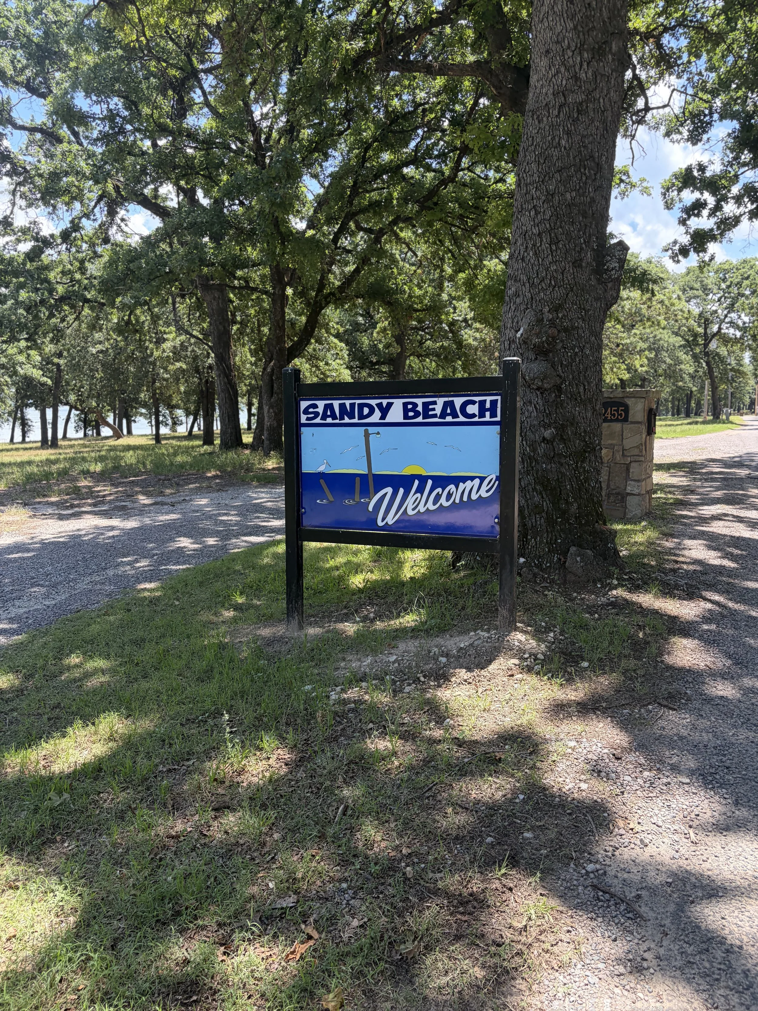 A beach sign reading 'Sandy Beach Welcome' in front of a large tree with green foliage, on a sunny day with clear skies and a gravel path nearby.