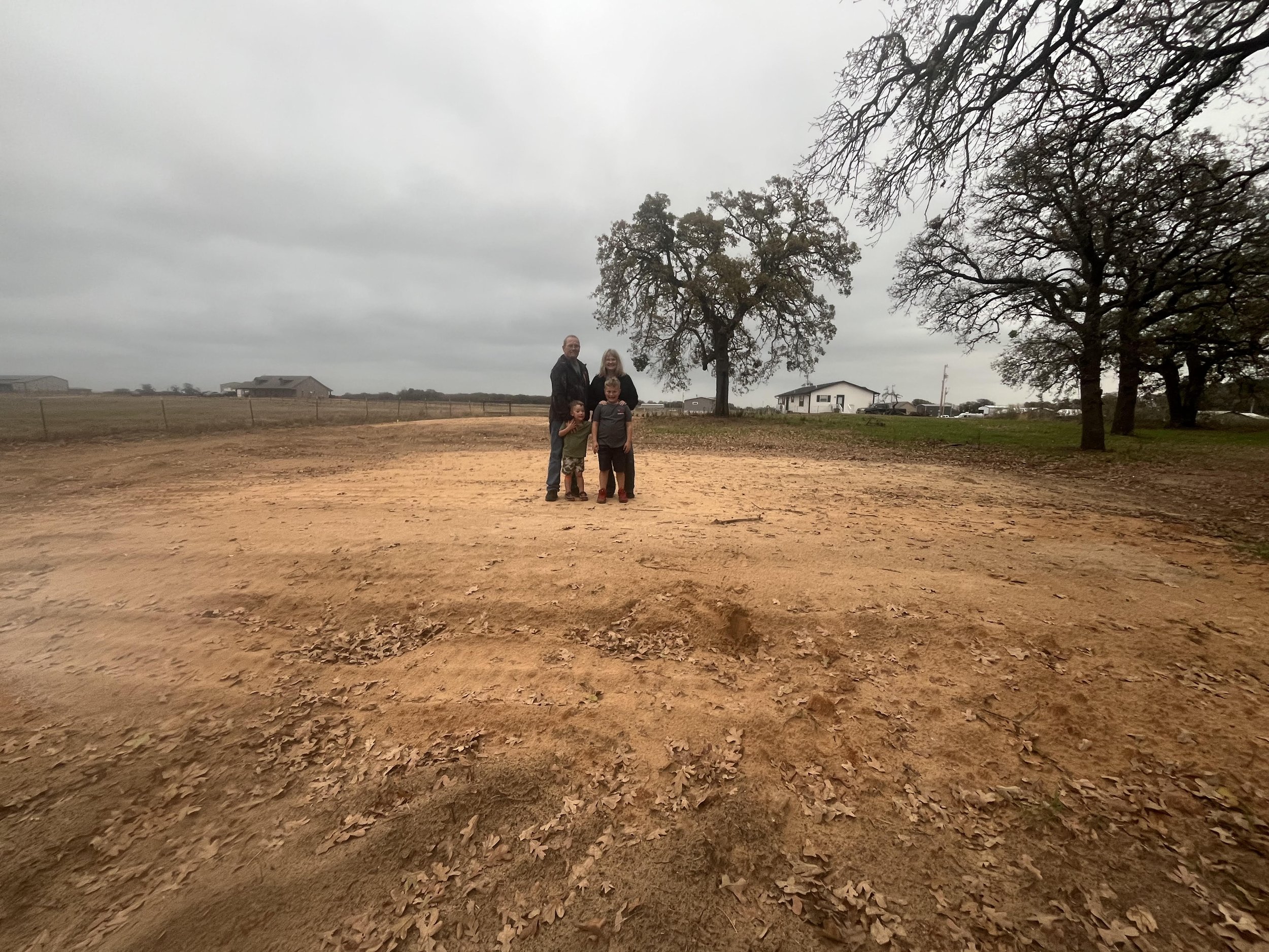 A man, woman, and two children standing on a dirt ground surrounded by leafless trees, with buildings and overcast sky in the background.