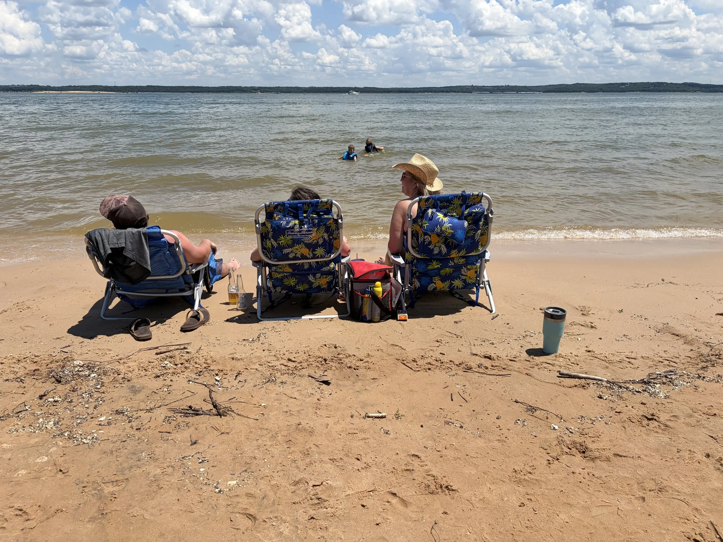 People relaxing on beach chairs on sandy shore, with two children playing in the water, and a woman wearing a wide-brimmed hat and sunglasses looking towards the water, under a partly cloudy sky.