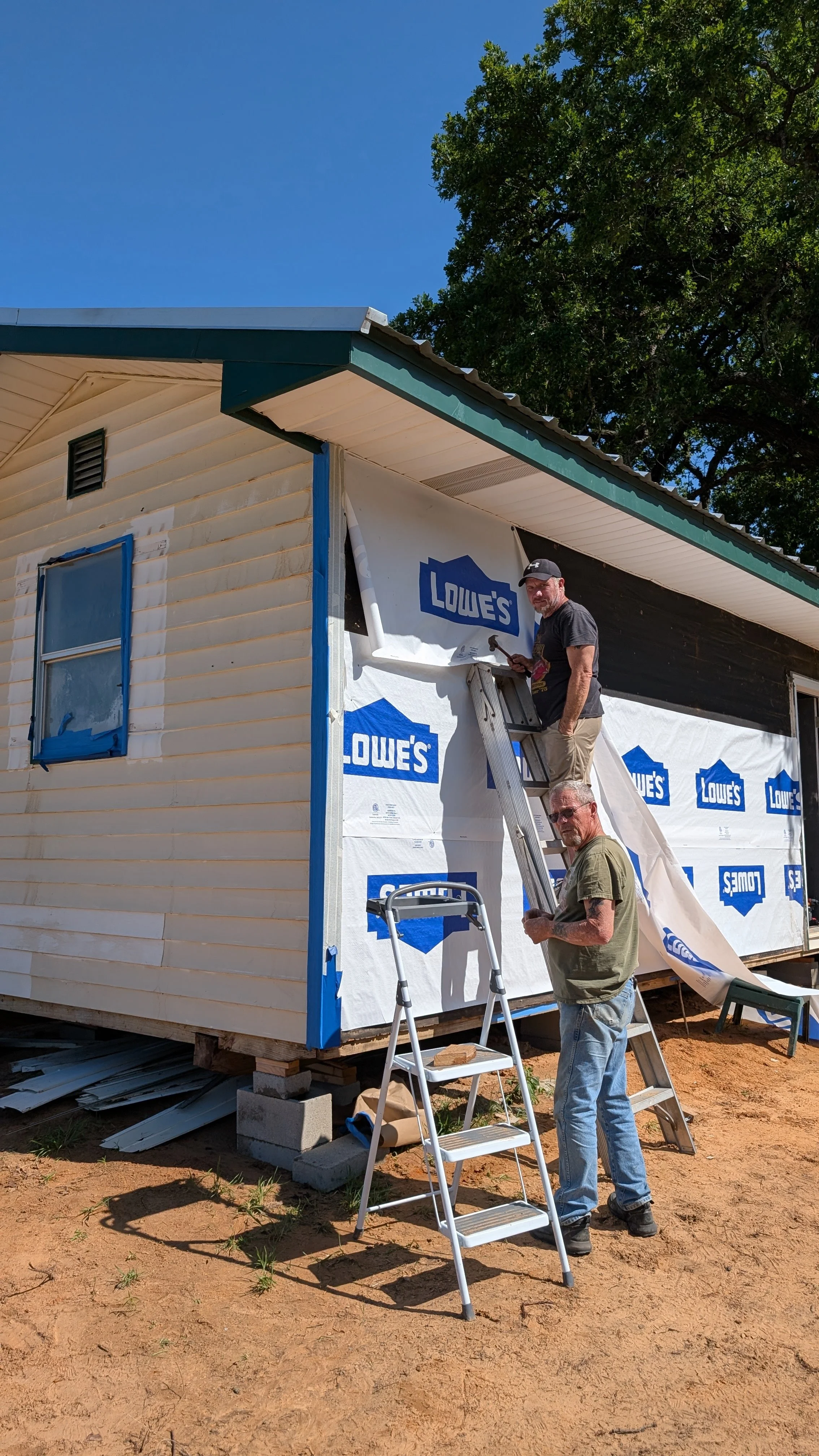 Two men working on the exterior of a house under construction, with house wrapped in Lowe's construction material, and ladders placed against the house, on a dirt ground with trees in the background.