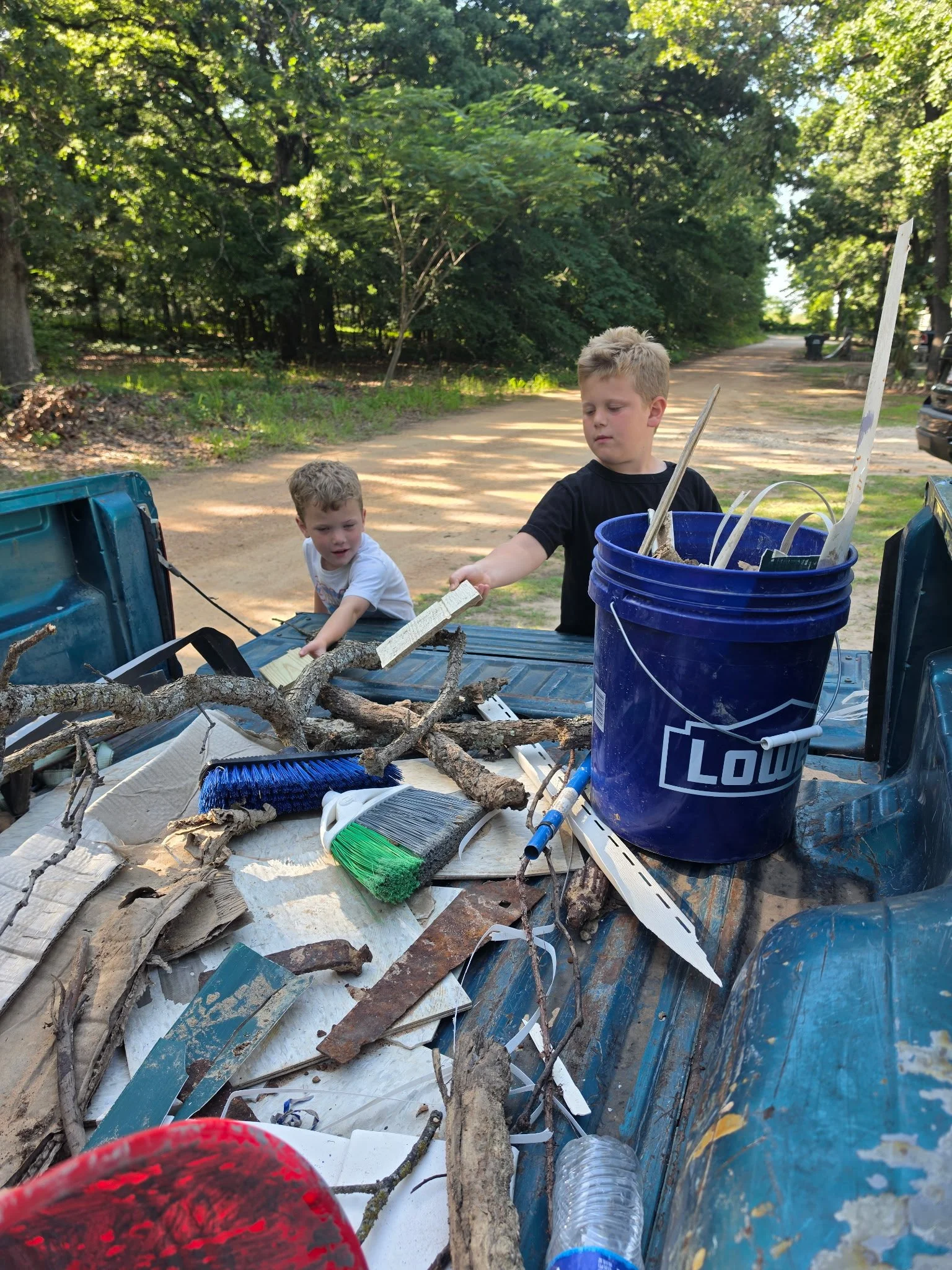 Two young boys, one in a black shirt and the other in a white shirt, are cleaning up trash and debris, including branches, plastic bottles, and other litter, in the back of a pickup truck on a dirt road surrounded by trees.