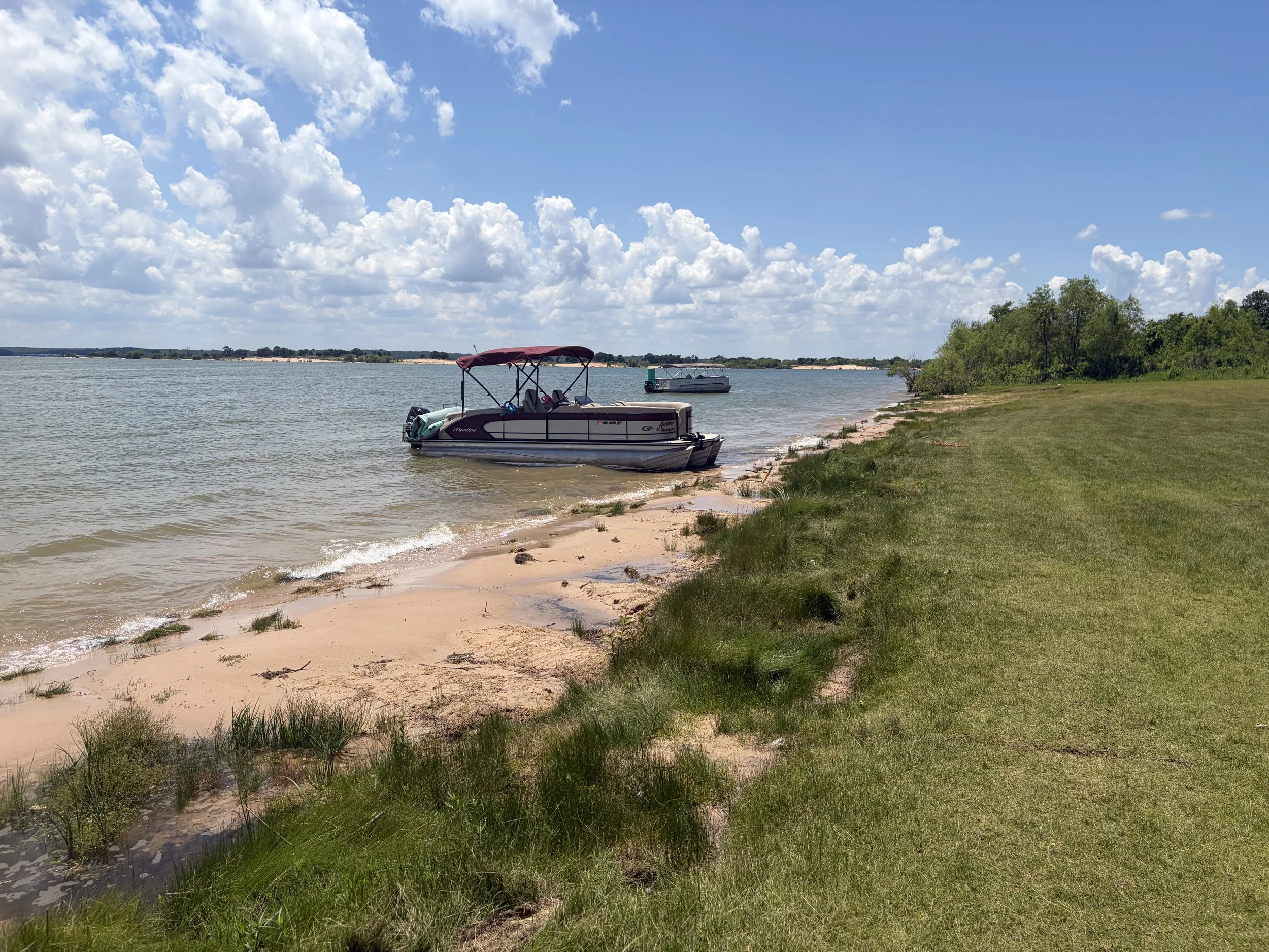 A sandy shoreline with grassy patches, two boats docked near the water, green trees in the background, and a blue sky with white clouds.