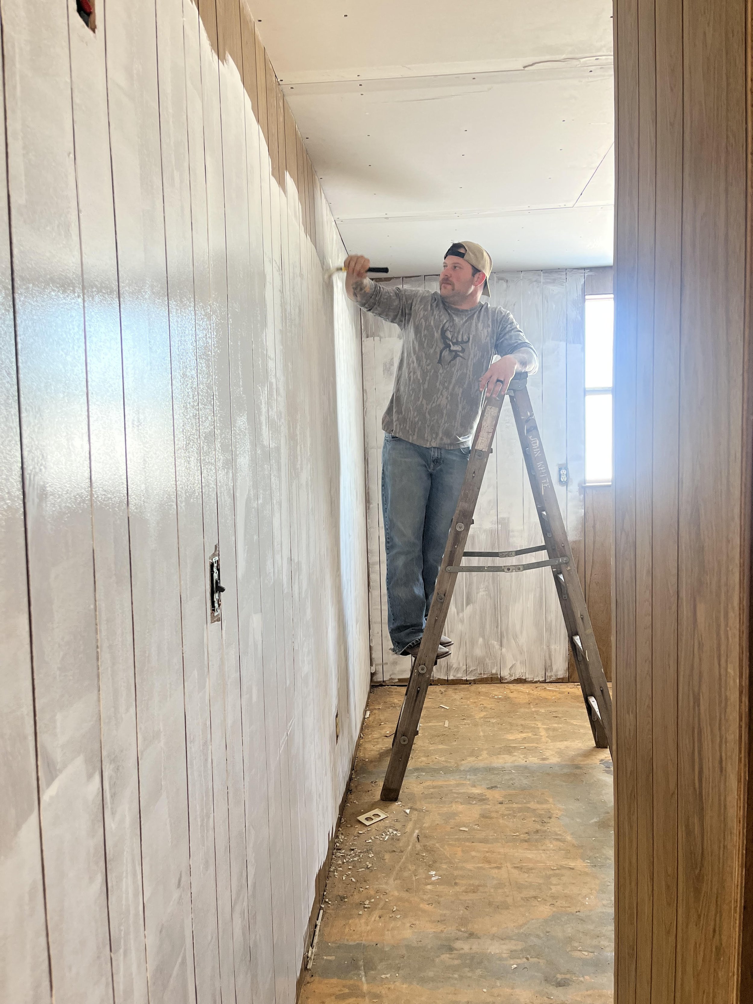 A man standing on a ladder painting a wall in a room that is under renovation. The room has wood paneling on one side and exposed drywall on the ceiling.