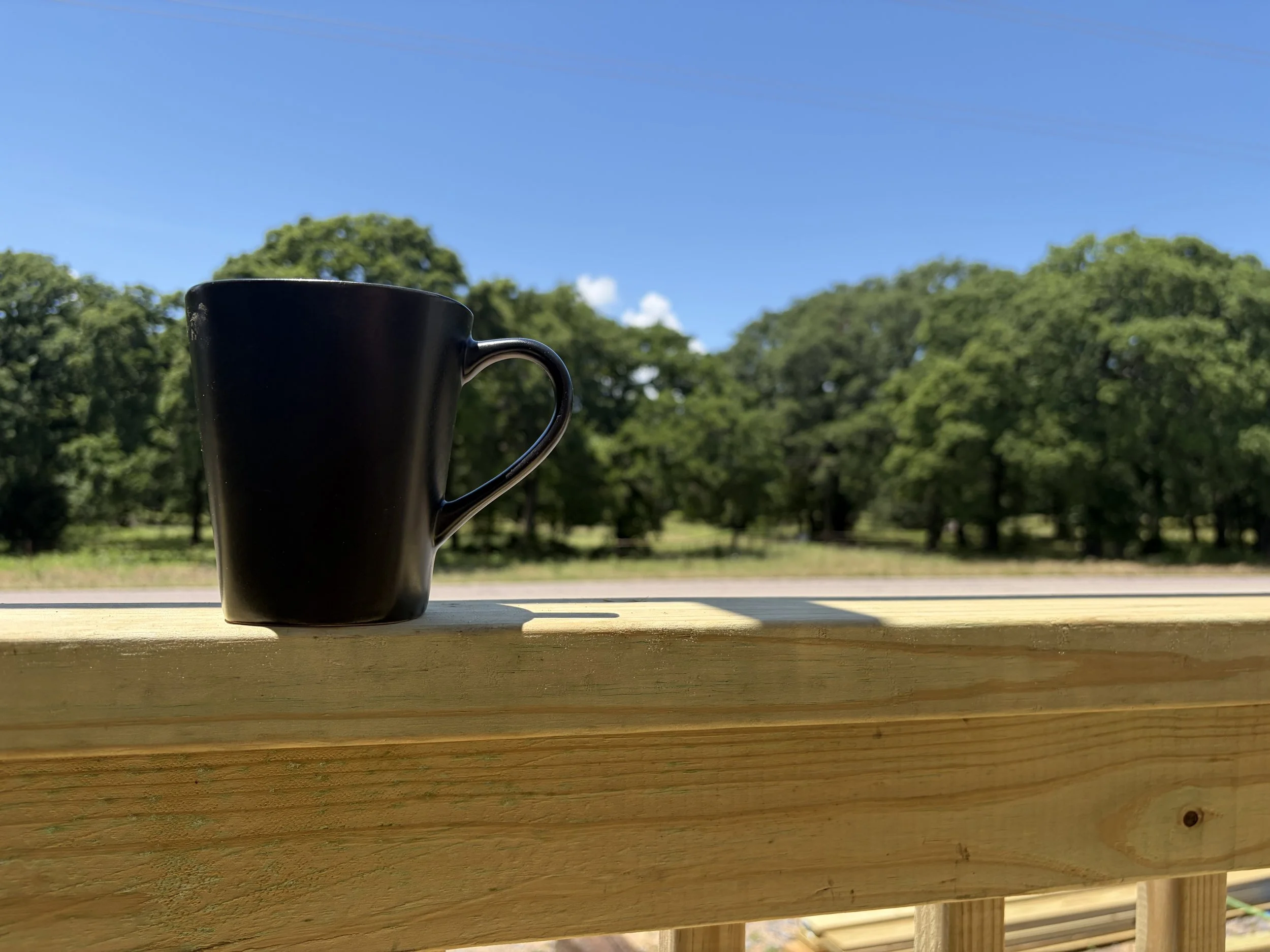 Black coffee mug on a wooden surface with a background of green trees and a bright blue sky.