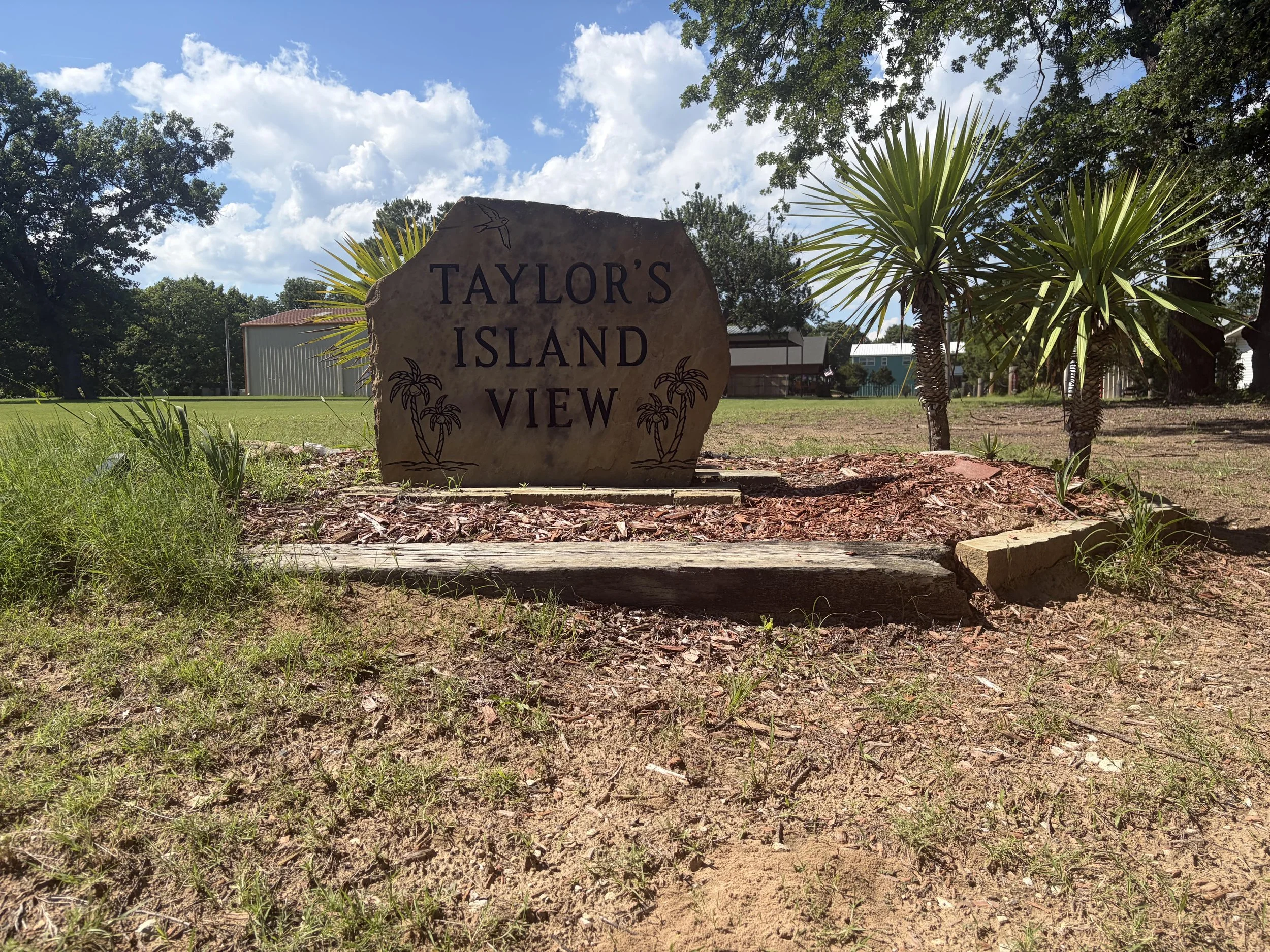 A large stone sign with the name "Taylor's Island View" in front of palm trees and a grassy area, with a blue sky and scattered clouds overhead.