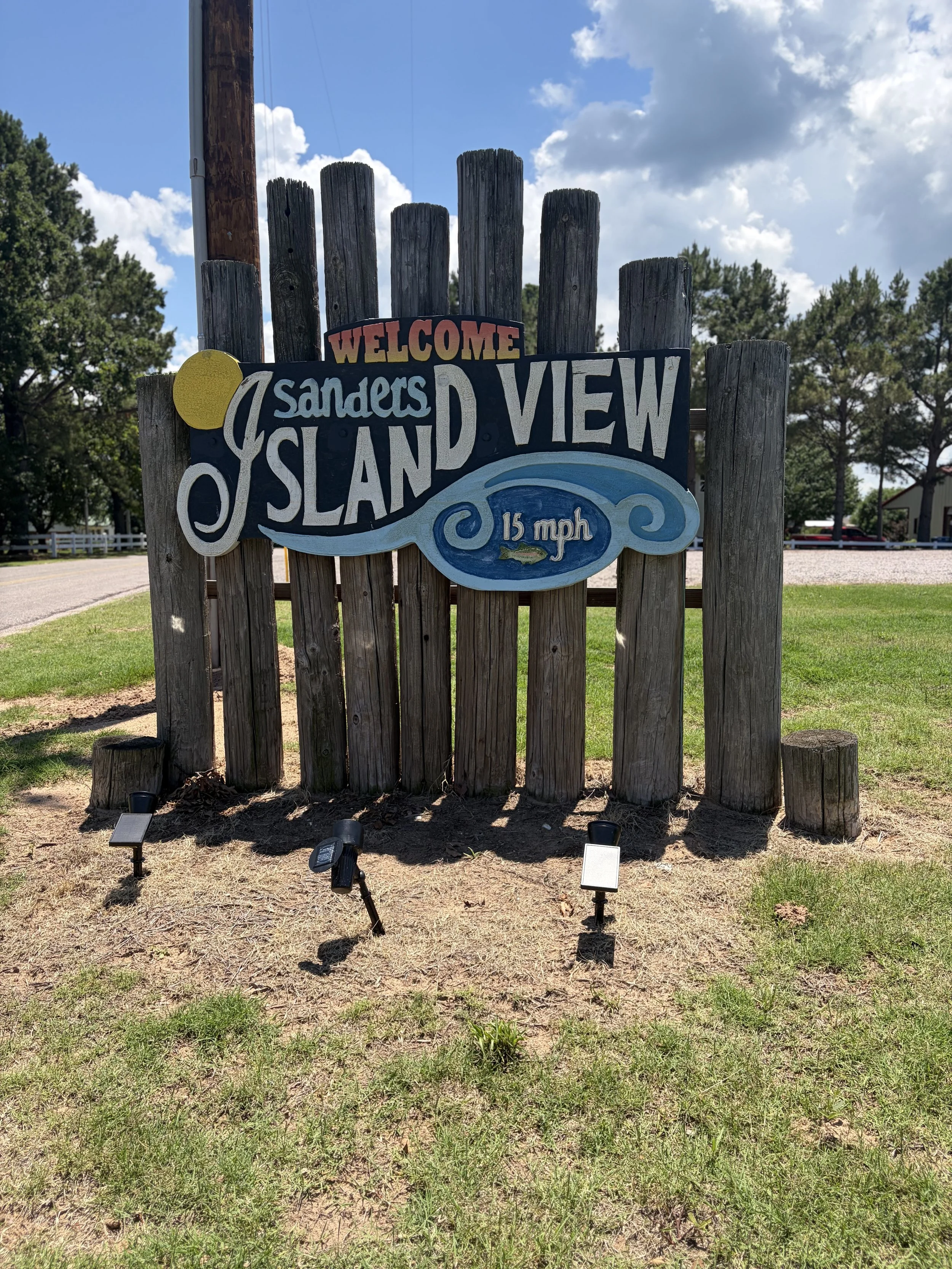 Wooden signboard with colorful text welcoming visitors to Sanders Island and advising 15 mph speed limit. The sign is mounted on a wooden fence near a grassy area with sunlight.