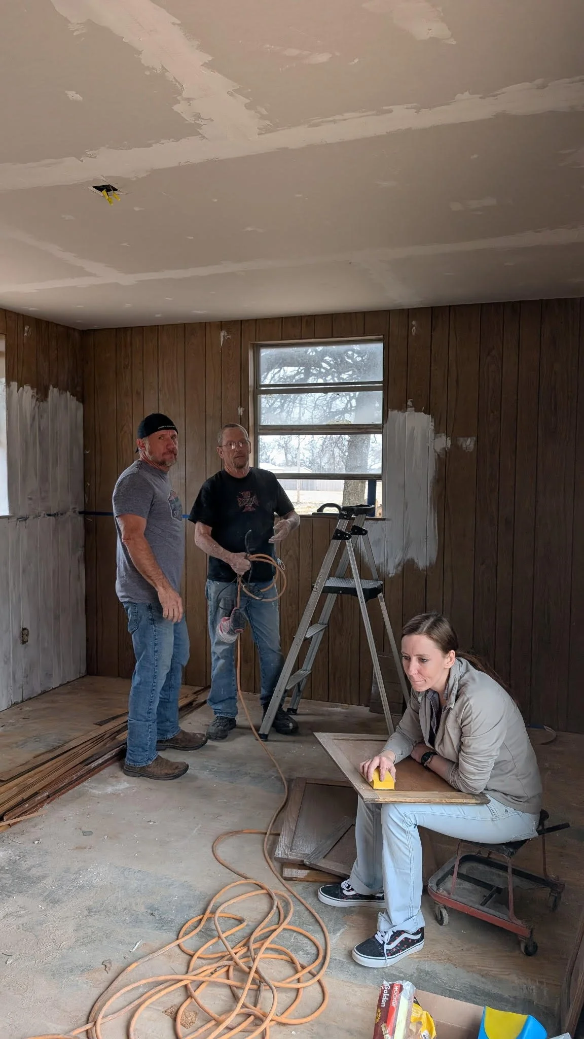 Three people working on renovation of a room with wood-paneled walls and ceiling. Two men are standing, one holding a cord, and a woman is seated, sanding a wooden surface. Tools and materials are scattered on the floor.