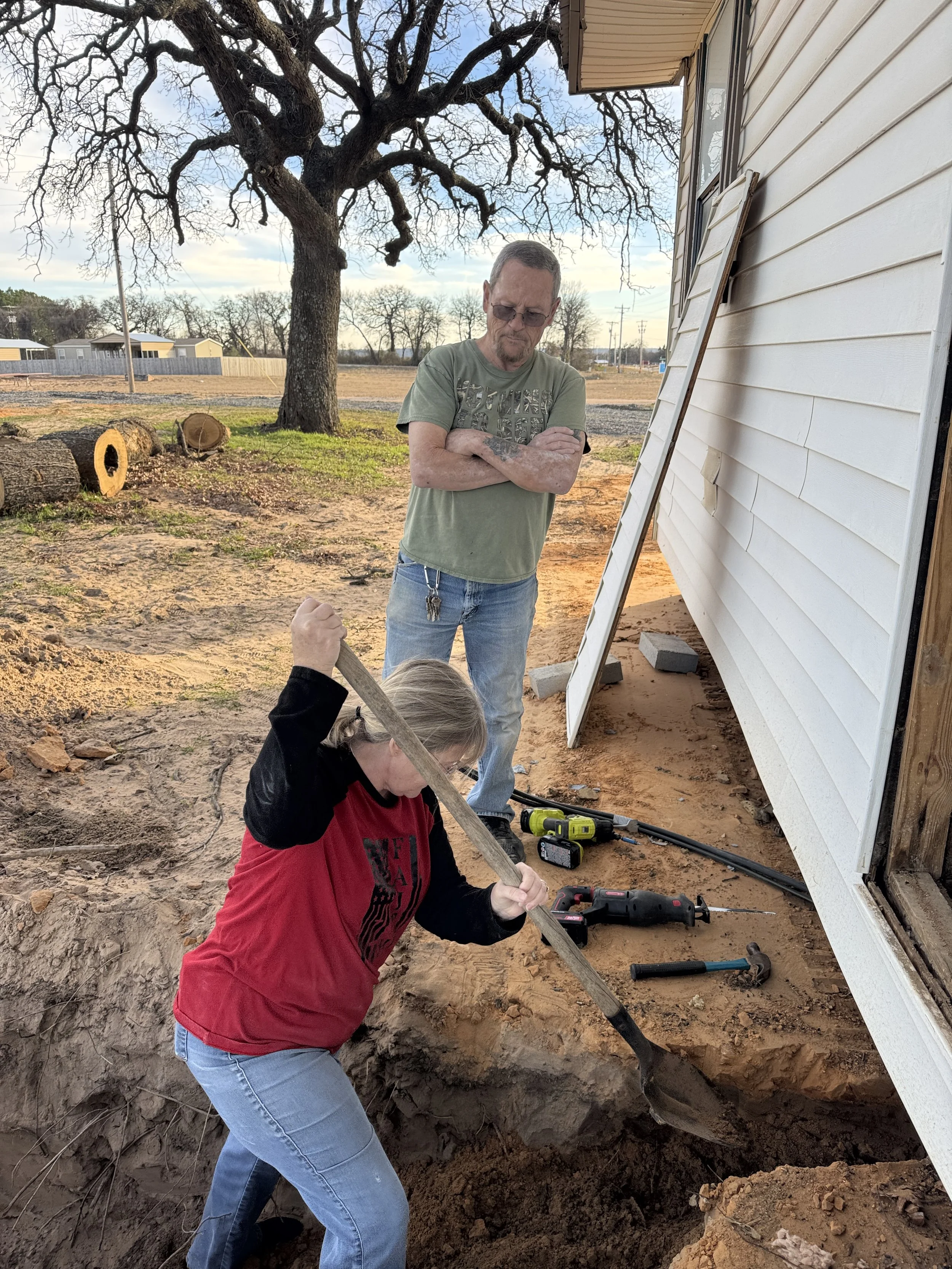 Two women and one man working on a construction project outside a house. The woman in the foreground is digging with a shovel, and the man in the background is standing with crossed arms. The house has white siding, and there is a large tree and logs in the yard.