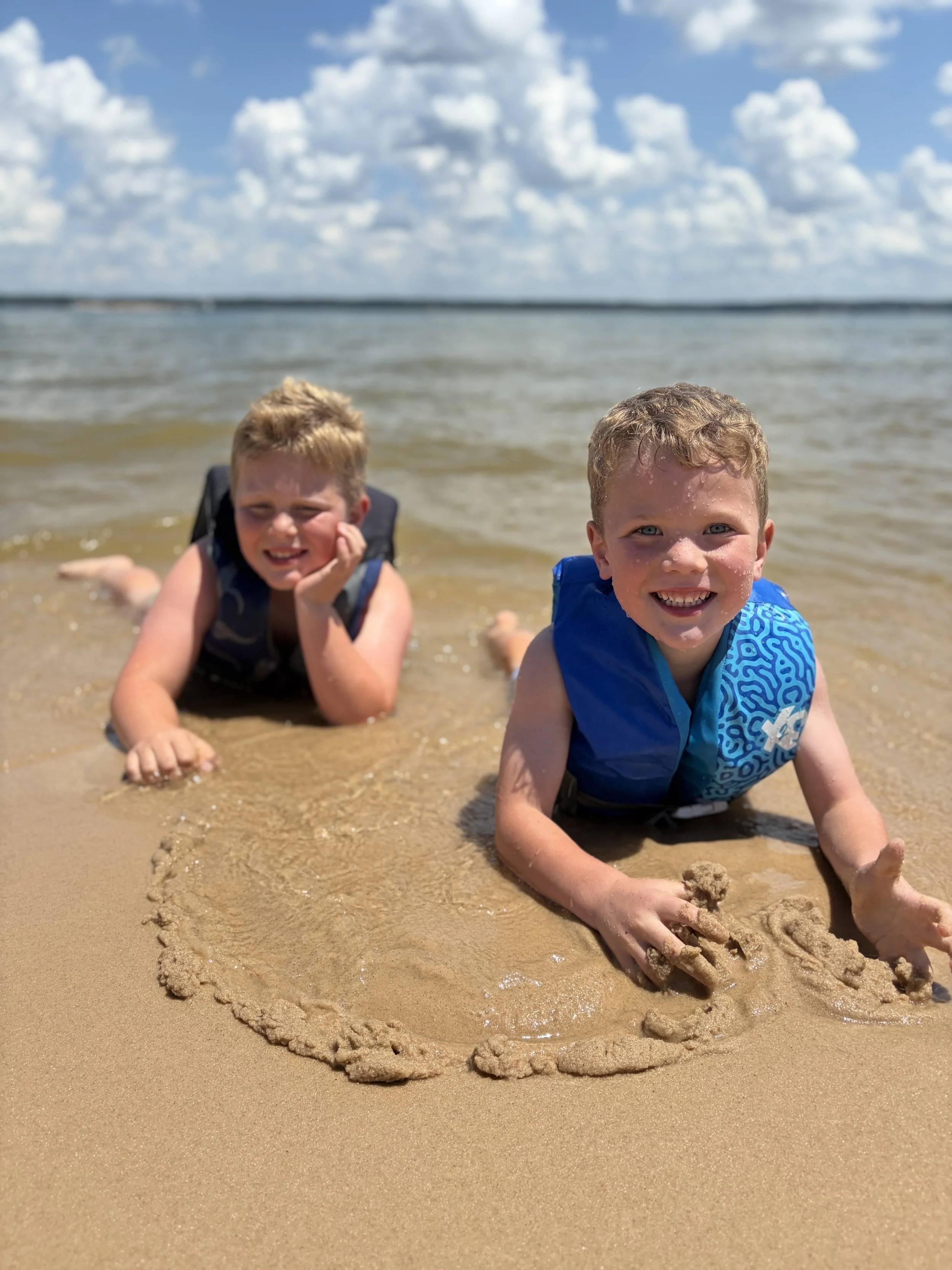 Two young boys playing with sand and making a heart shape on the beach during sunny weather, with the ocean and cloudy sky in the background.