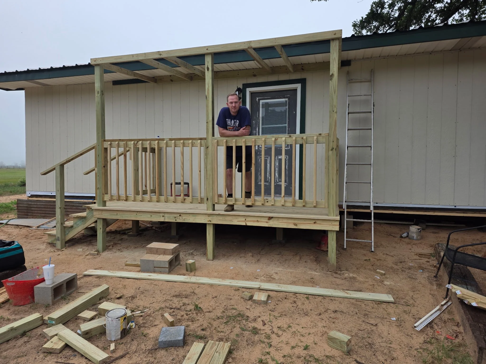 Man standing on a wooden porch under construction in front of a house, with tools and building materials scattered around.