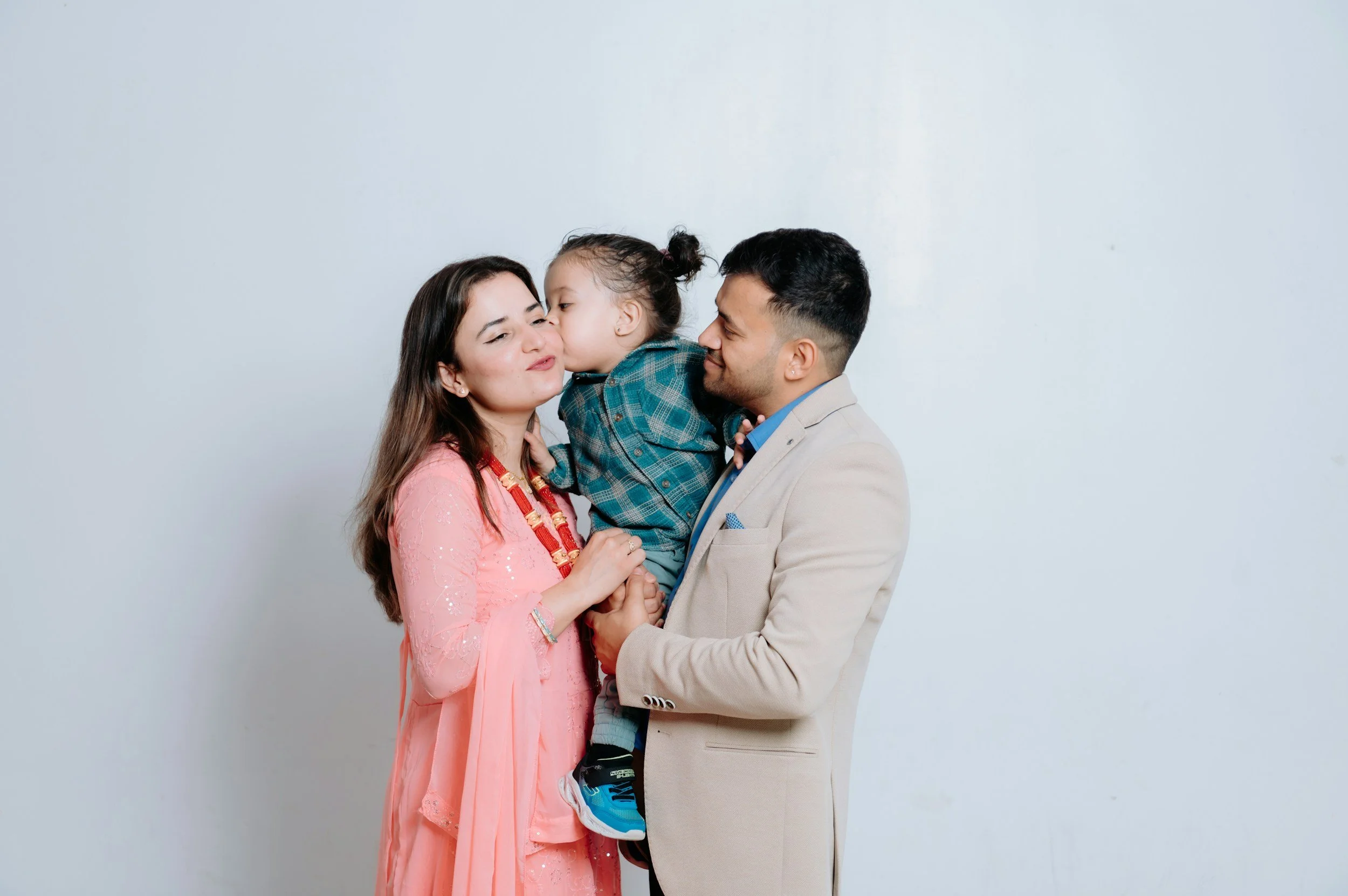 A family of three enjoying a moment together against a plain background. The woman is wearing a pink traditional dress with jewelry, the man is in a beige suit jacket, and their young son is kissing the woman's cheek.
