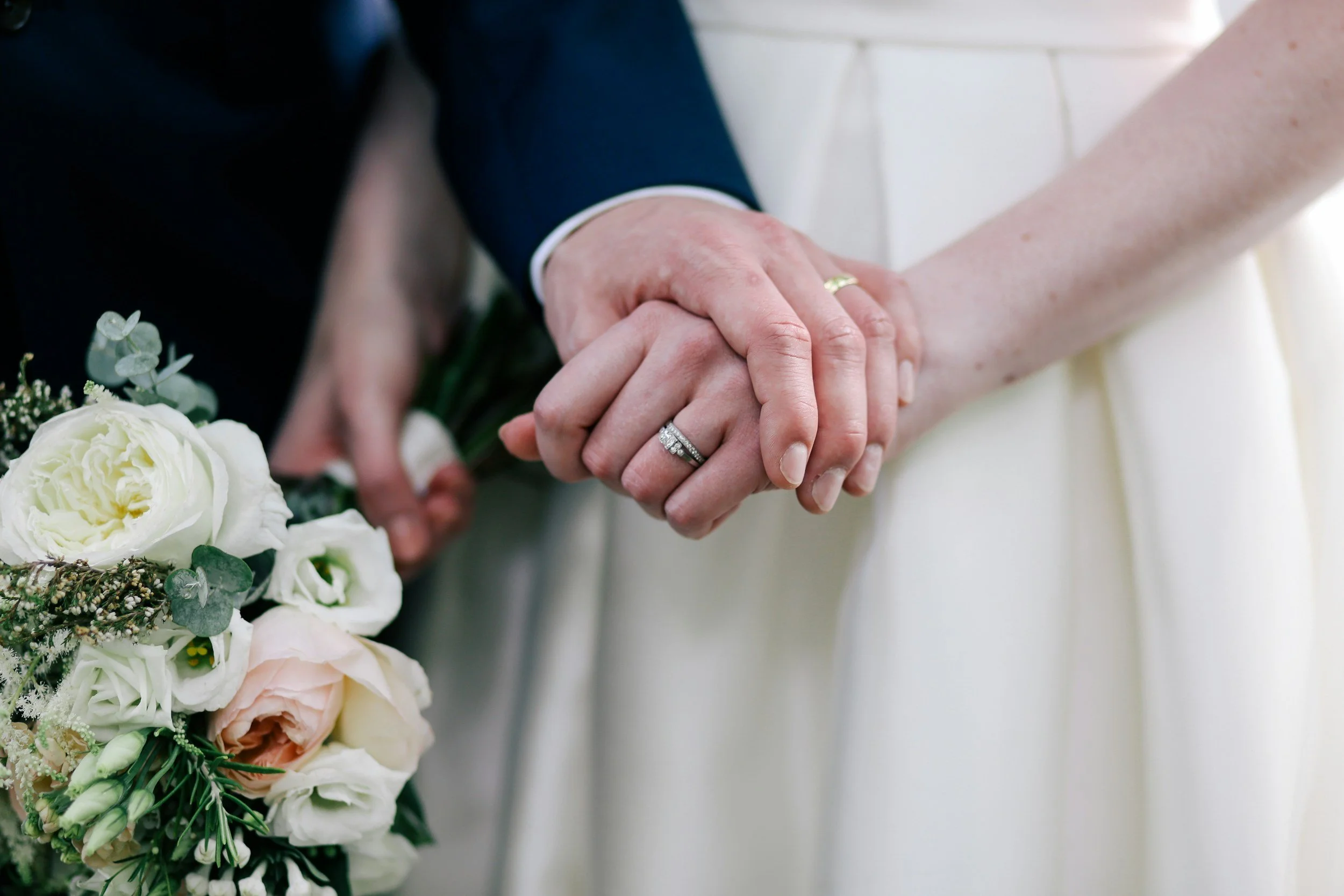 Close-up of a bride and groom holding hands, showing wedding rings, with a bouquet of white and pale pink flowers in the foreground.