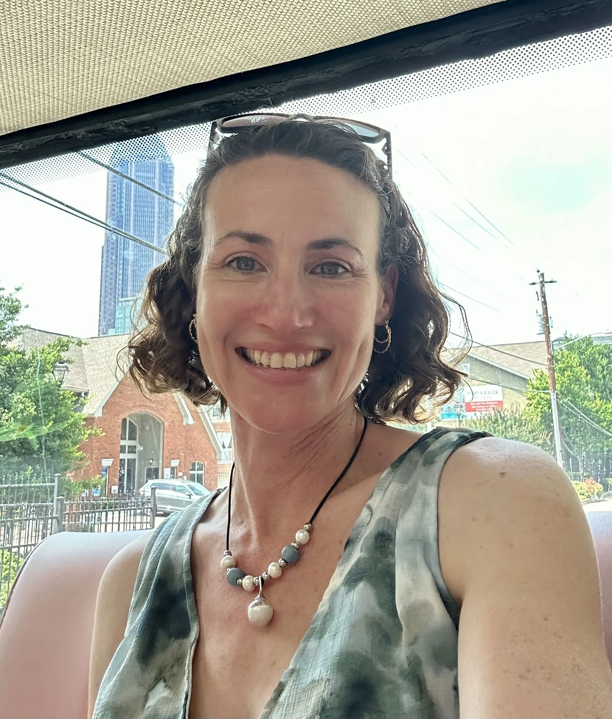 A woman smiling outdoors in front of a cityscape with tall buildings, trees, and power lines, wearing a sleeveless top, a beaded necklace, and sunglasses on her head.