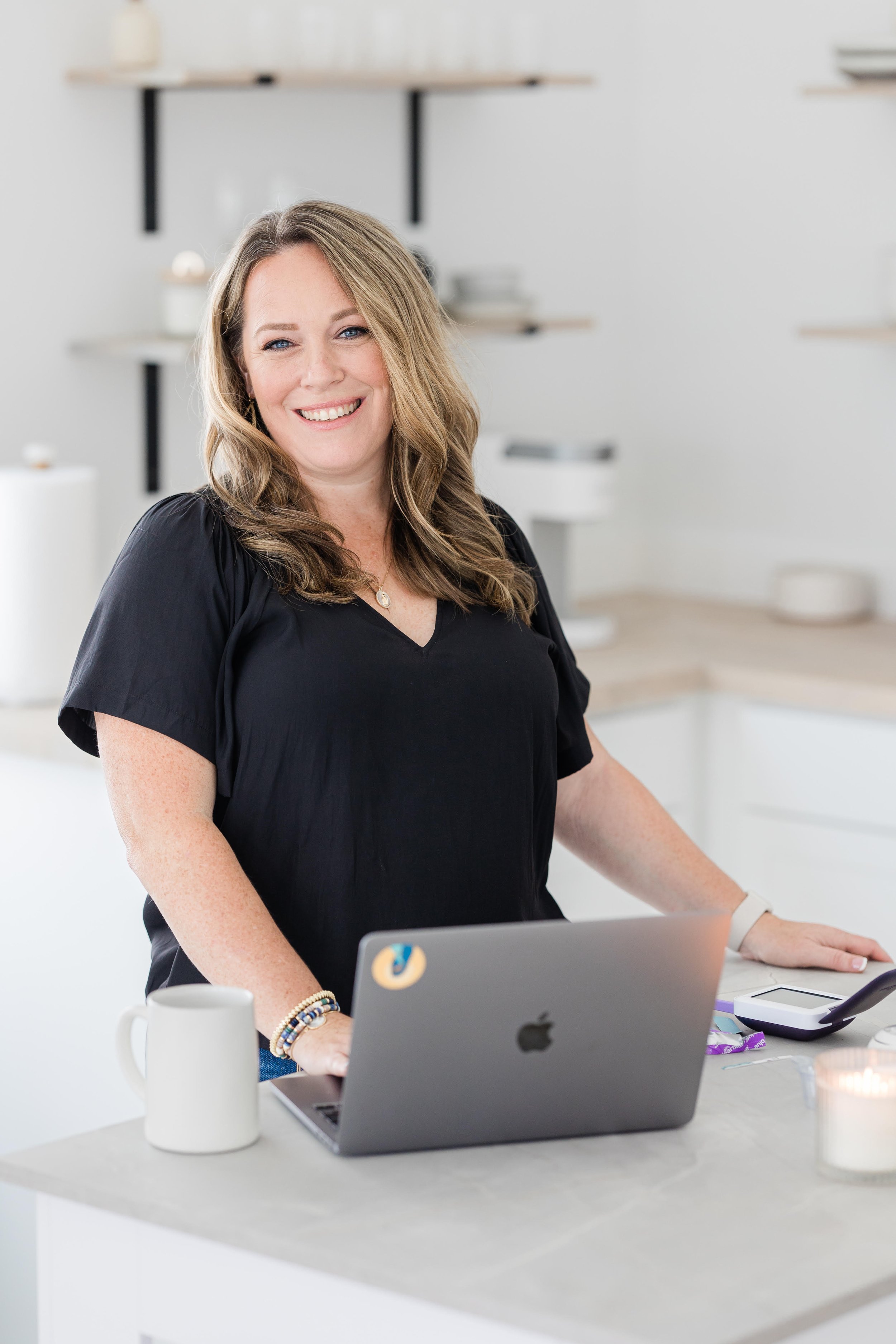 A woman with shoulder-length wavy hair and wearing a black top smiling at a workspace. She has a bracelet on her right wrist and is sitting in front of a laptop with an Apple logo. There is a white mug, a smartphone, and a few snacks on the table. The background shows white shelves with minimal decor.