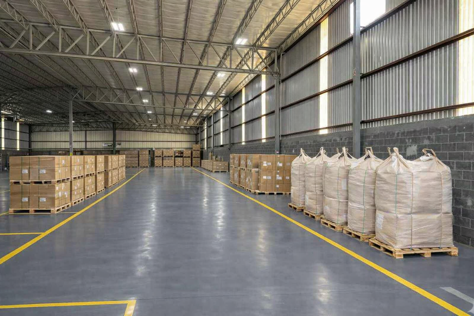 Almacén industrial con cajas y sacos de productos en pallets, con paredes de metal y piso gris, iluminado con luces de techo.