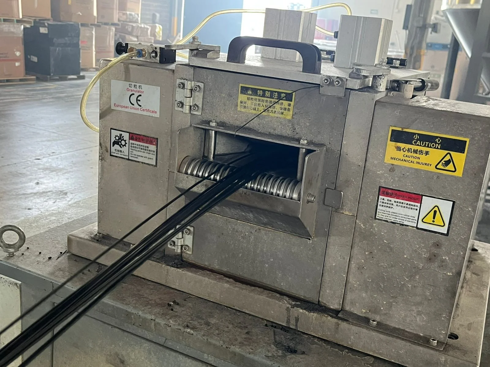 Máquina industrial de tamaño mediano con etiquetas en chino y en inglés, en un entorno de fábrica, con cables negros saliendo de ella y advertencias de seguridad en la parte superior y lateral.