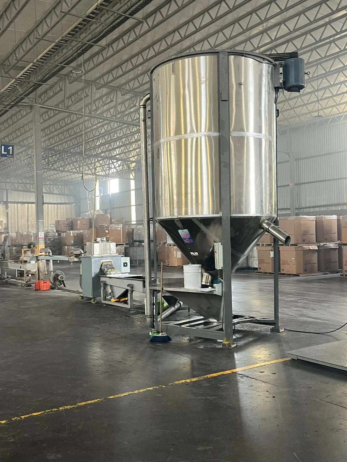 Tanque de acero inoxidable en una fábrica, con maquinaria y cajas en el fondo, en un ambiente industrial.