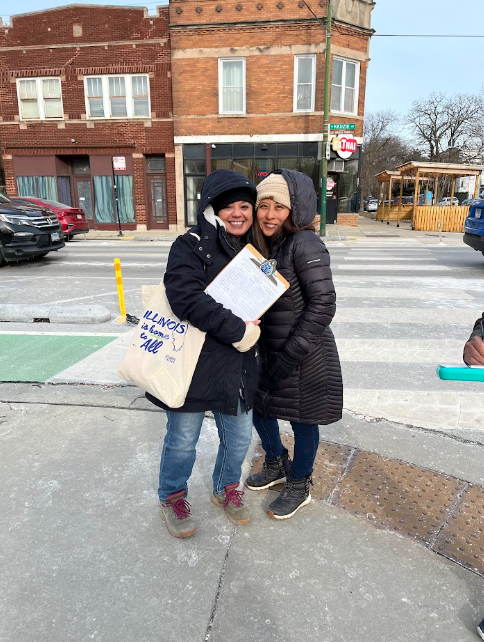 Two women standing on a city sidewalk, smiling, with one holding a clipboard, dressed warmly in jackets and hats, in front of a street with parked cars, brick buildings, and a corner store.