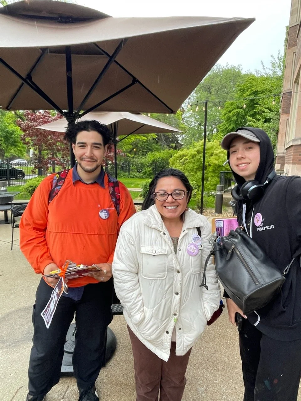Three people standing outdoors under umbrellas, smiling at the camera. One man is holding pamphlets, and the others are women wearing badges. There are trees and a building in the background.
