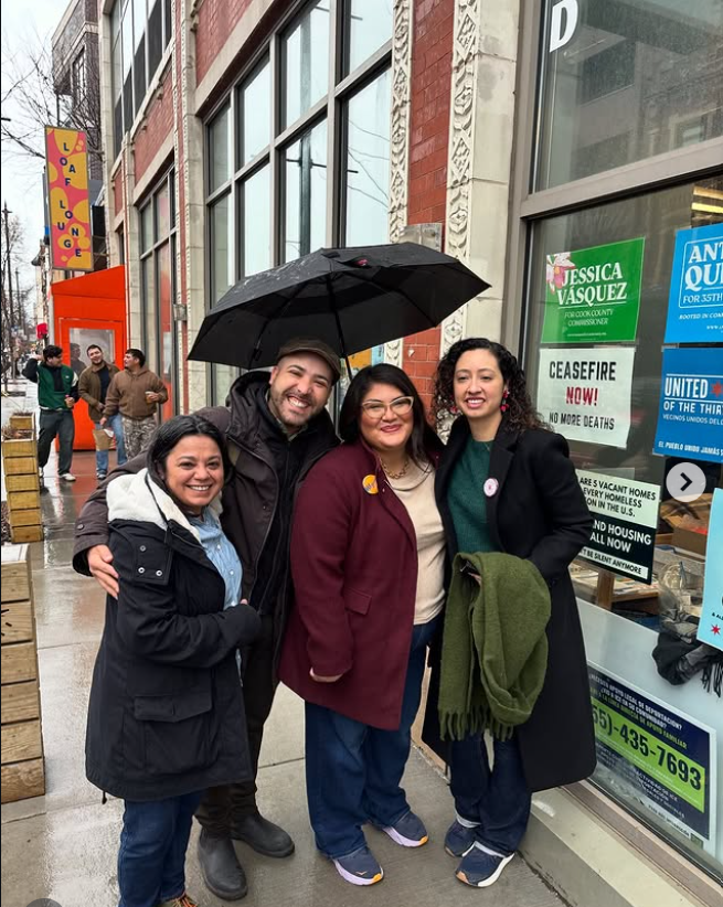 Four people standing outside on a rainy day in front of a building with signs, smiling, with one holding an umbrella over them.
