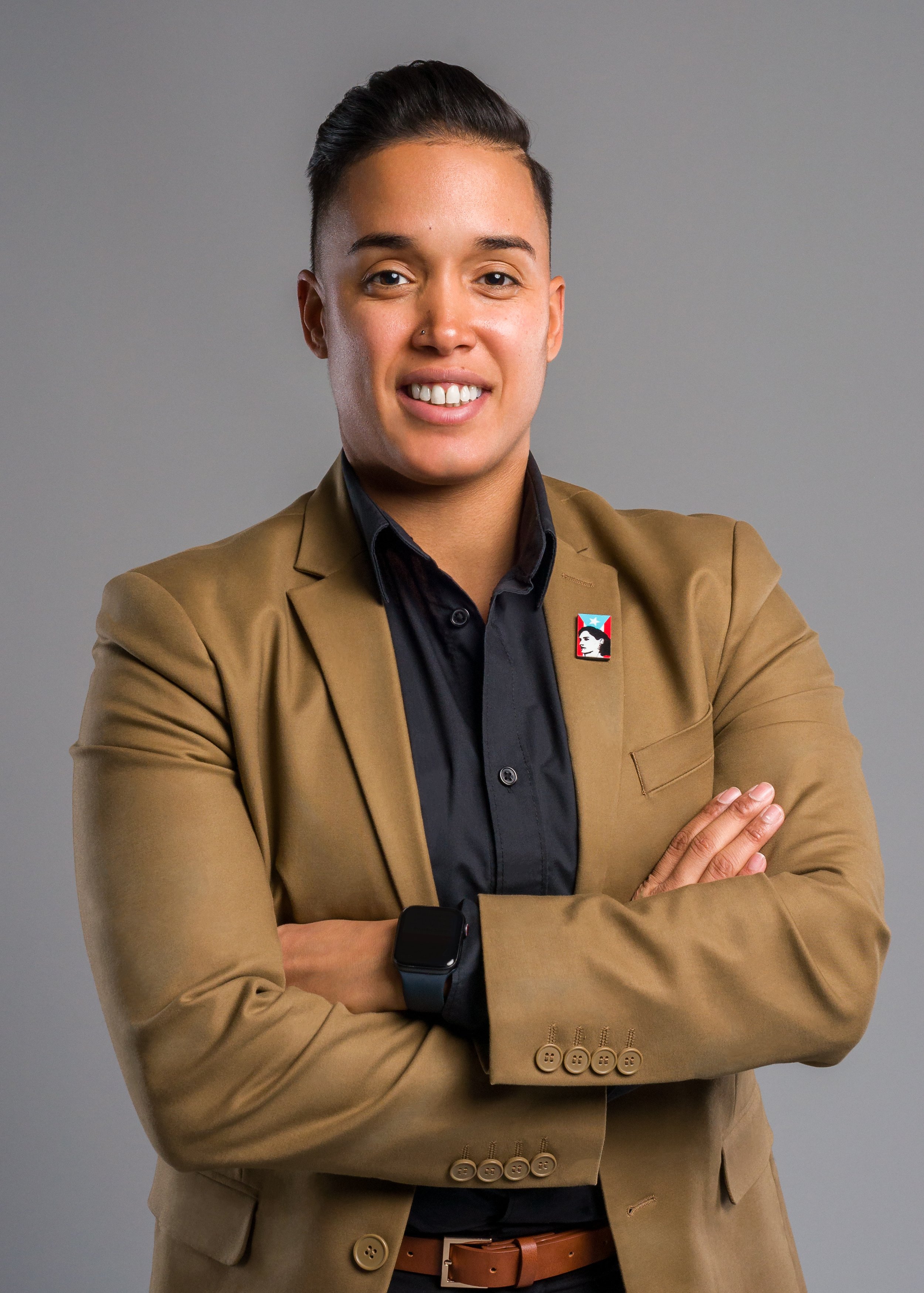A person with short black hair, dressed in a tan blazer with buttons on the sleeves, black shirt, brown belt, and wearing a smartwatch, standing with arms crossed against a plain gray background.