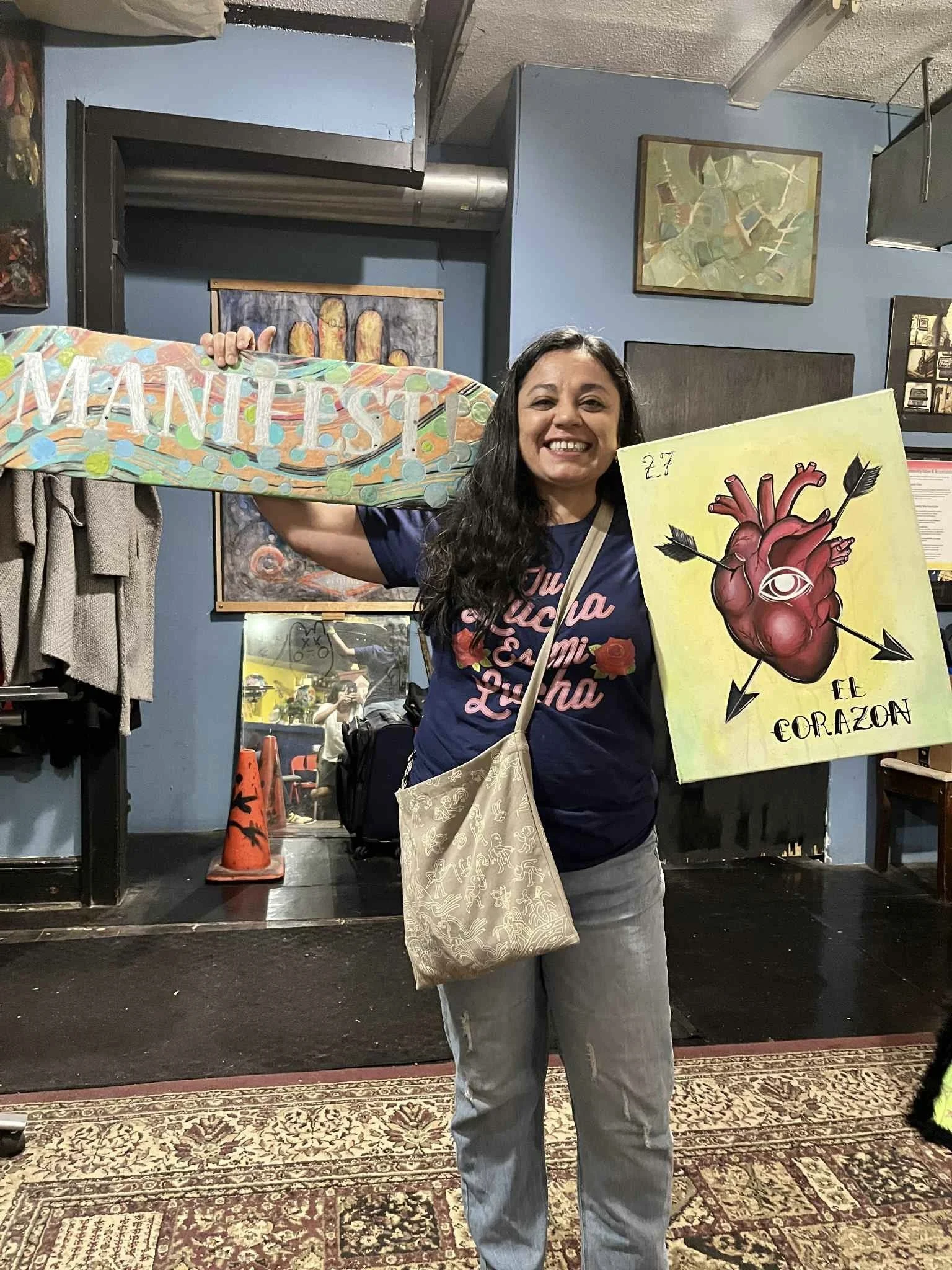 A woman with long dark hair smiling, holding a colorful sign with the words "MANI PILS" and standing in an indoor art gallery or studio. She is wearing a blue T-shirt with pink lettering and has a beige shoulder bag. The background shows artwork on the walls, a mirror, and a person taking her picture in the reflection.