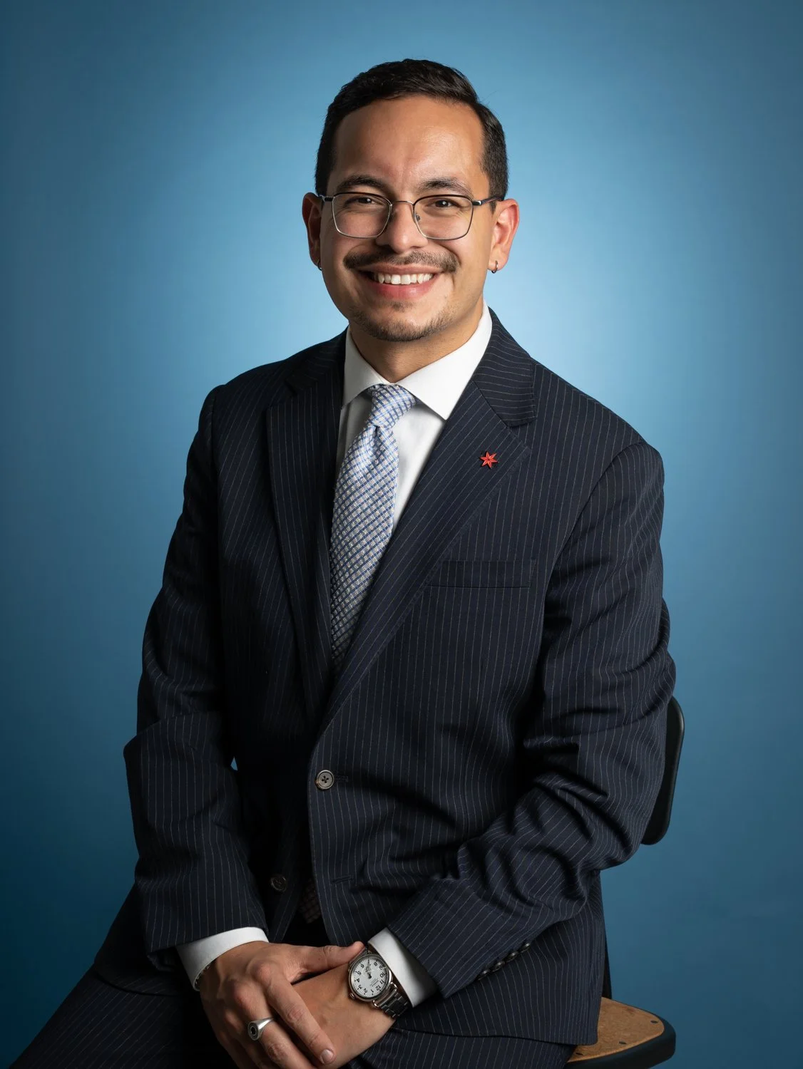 A smiling man in a navy pinstripe suit, light blue tie, and glasses, sitting against a blue background.