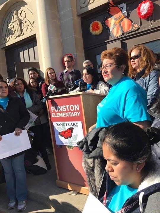 Group of people gathered outside Funston Elementary School for a press conference or event, with a woman speaking at a podium decorated with a butterfly and sanctuary logo, and others listening or holding signs.