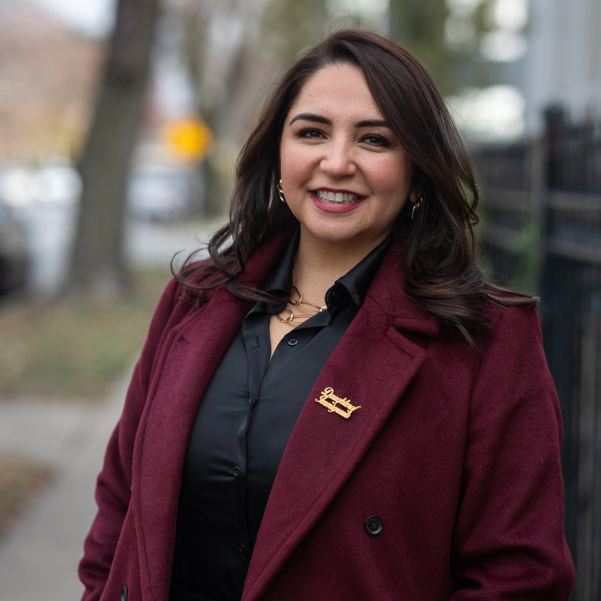 Close-up of a smiling woman with dark hair and earrings, wearing a maroon coat over a black shirt, outdoors with blurred trees and a yellow traffic sign in the background.