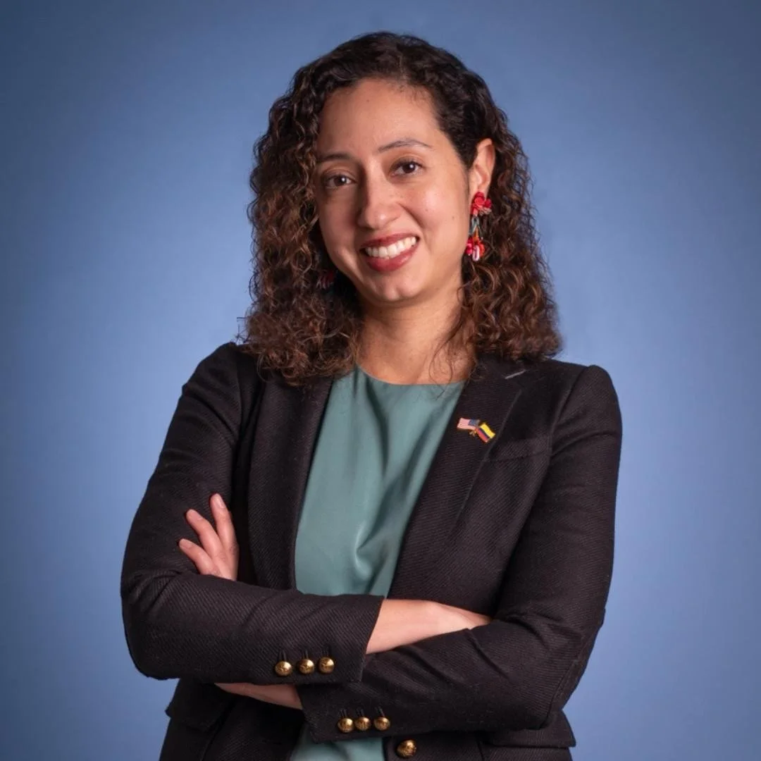 A woman with curly brown hair, wearing a black blazer with gold buttons, a teal blouse, red floral earrings, and a pin with American and German flags, standing with arms crossed against a blue background.