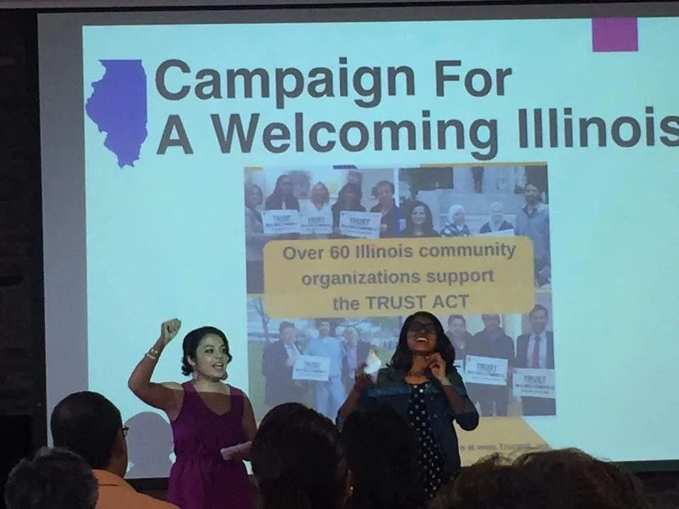 Presentation slide for a campaign titled 'Campaign For A Welcoming Illinois,' supported by over 60 Illinois community organizations advocating for the TRUST ACT, with a photo of protesters holding signs, and two women speaking in front of the slide.