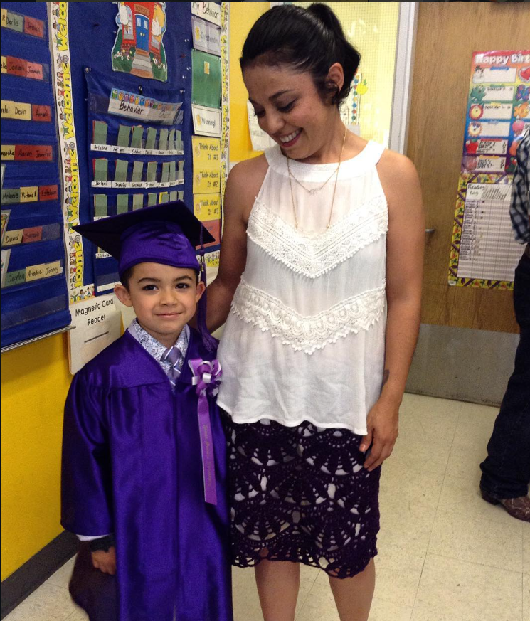 A young boy in a purple graduation cap and gown standing beside a woman in a white sleeveless blouse and a black lace skirt, at a school or classroom setting, celebrating a graduation event.