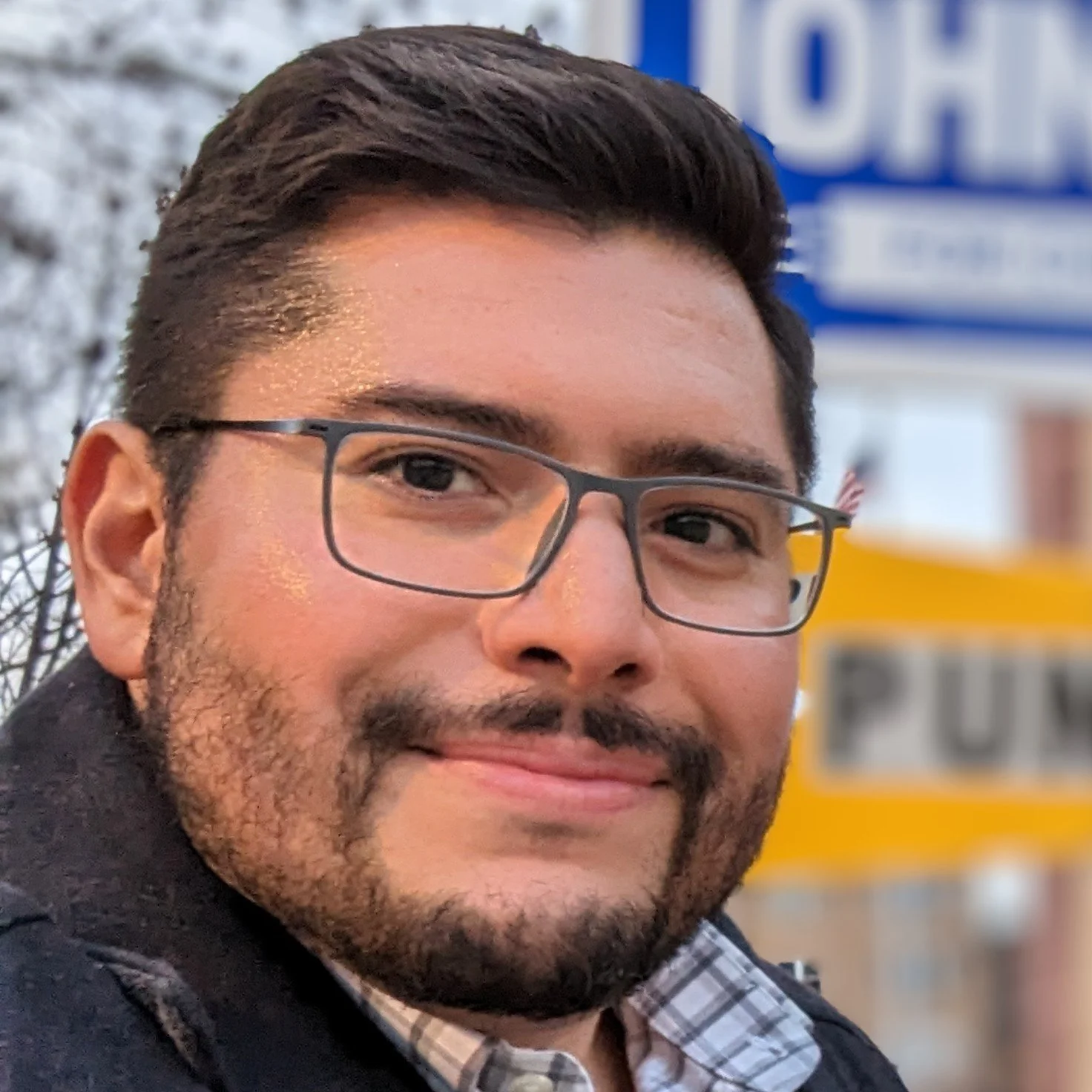 Close-up of a smiling man with glasses, dark hair, and a beard, outdoors near a blurry yellow and blue sign.