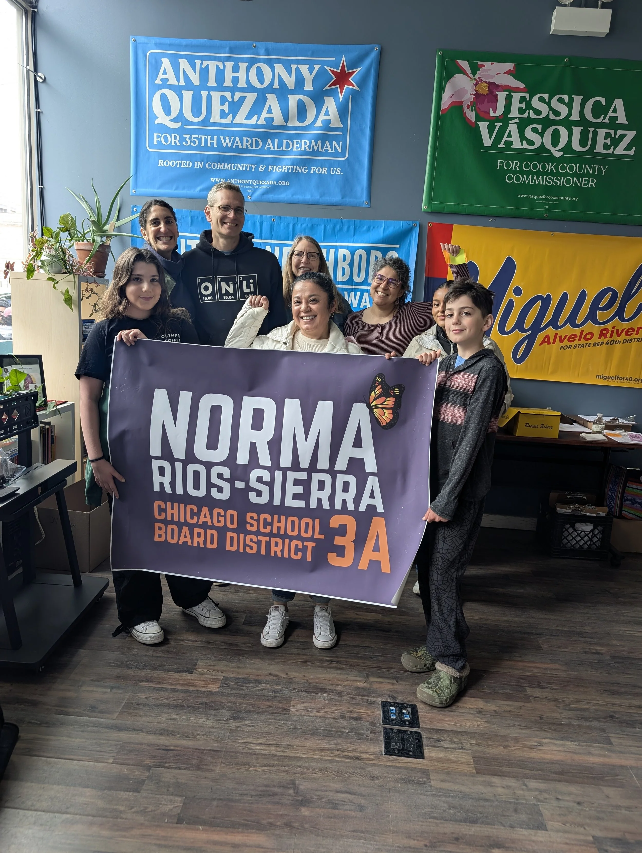 A group of six people standing indoors, holding a purple banner that reads 'NORMAND RIOS-SIERA CHICAGO SCHOOL BOARD DISTRICT 3A'. Behind them are several campaign banners on the wall, including supporters for Anthony Quezada and Jessica Vasquez. The group is smiling and posing for the photo, with some raising their fists.