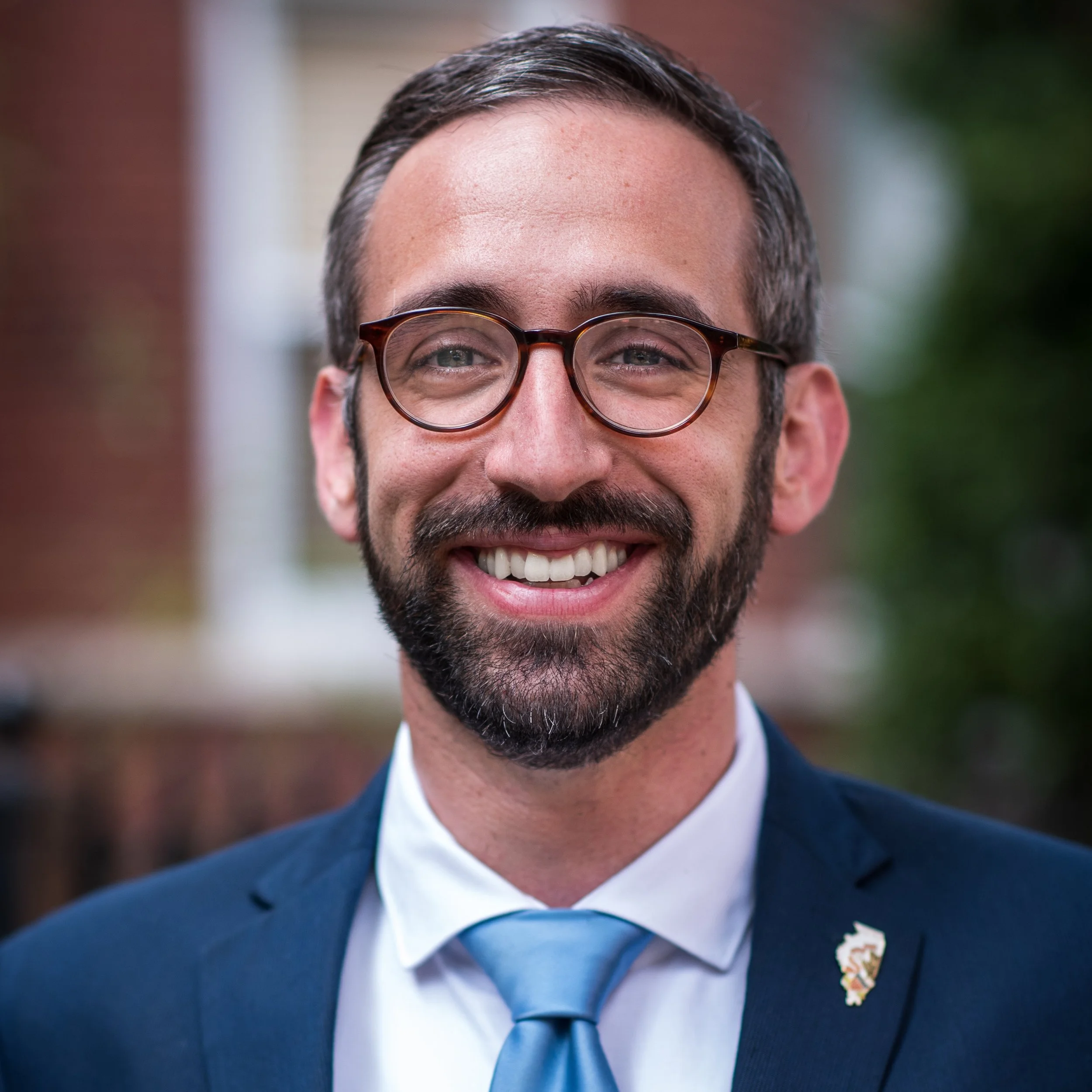 Close-up of a smiling man wearing glasses and a blue suit with a blue tie, outdoors, with blurred background.