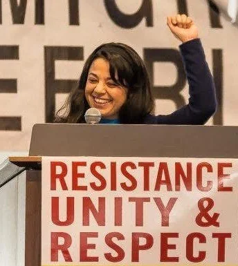 A woman at a podium with a sign that reads 'Resilience, Unity & Respect,' raising her fist in the air and smiling.