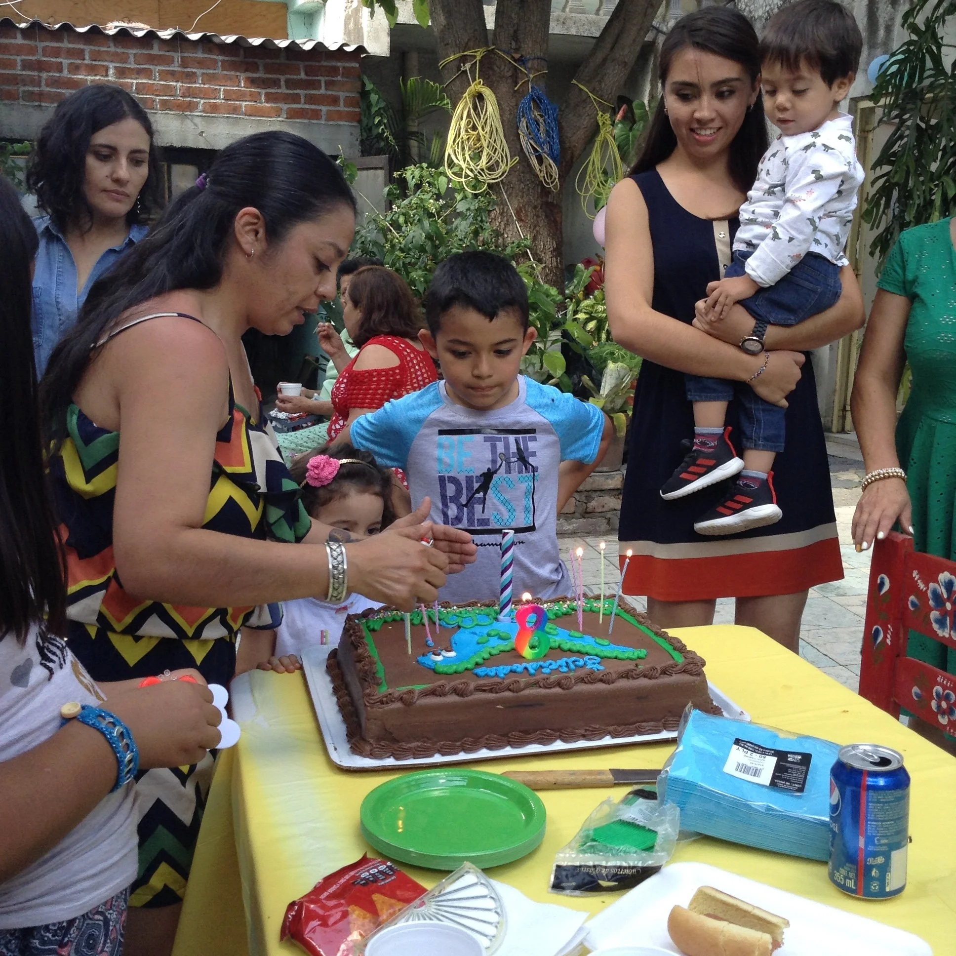 Children and adults gathered around a birthday cake with candles, decorations, and presents on a yellow table outdoors.