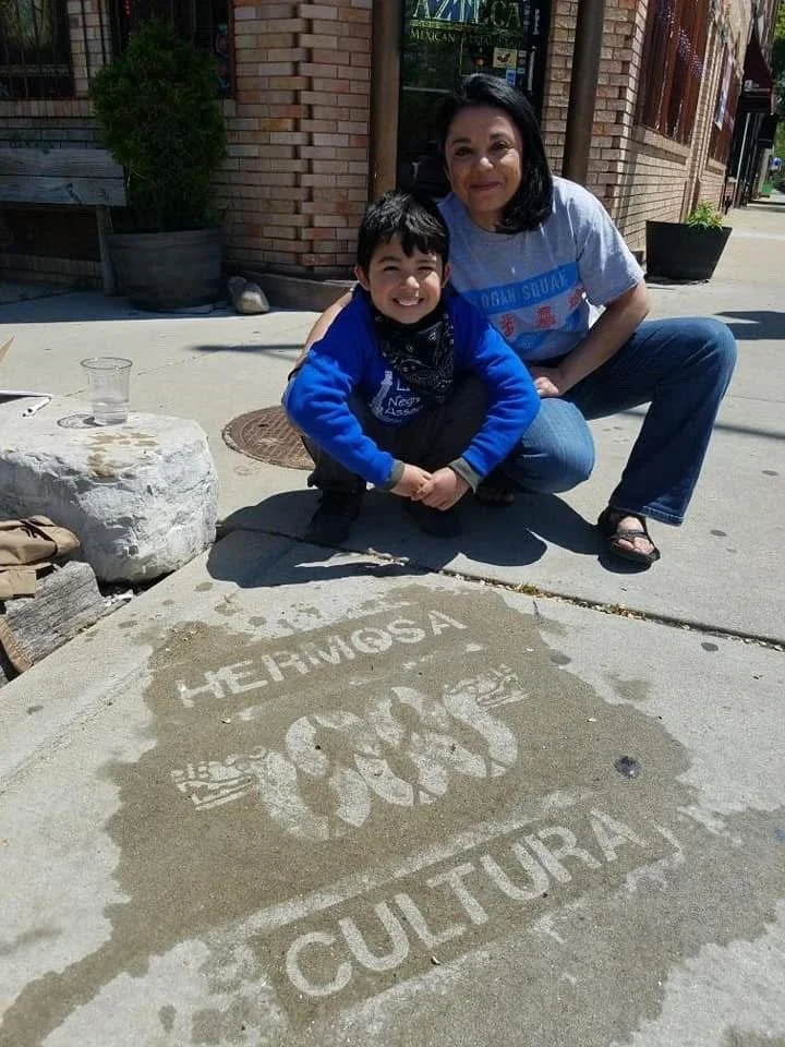 A woman and a young boy squatting on a sidewalk next to a sidewalk art of a bear paw print with the words 'Hermosa' above it and 'Cultural' below it, created with wet cement.