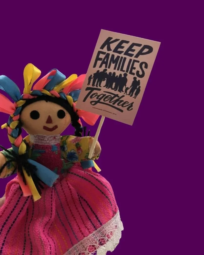 A handmade doll with colorful yarn hair, wearing a pink dress and holding a sign that reads 'Keep Families Together' with silhouettes of diverse people.