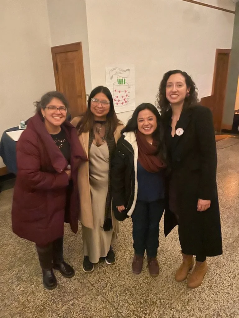 Four women standing indoors, smiling for a photo, with a wall and a poster behind them.