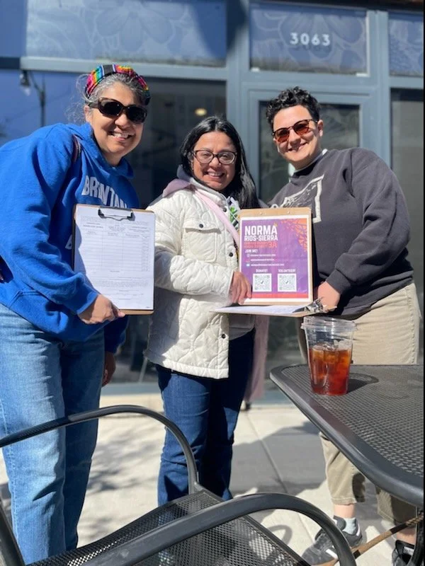 Three women standing outside a building, holding a framed certificate or award, smiling at the camera. One woman wearing sunglasses and a blue hoodie, another in a white coat, and the third in a dark gray sweatshirt. A table with a drink is visible in the foreground.