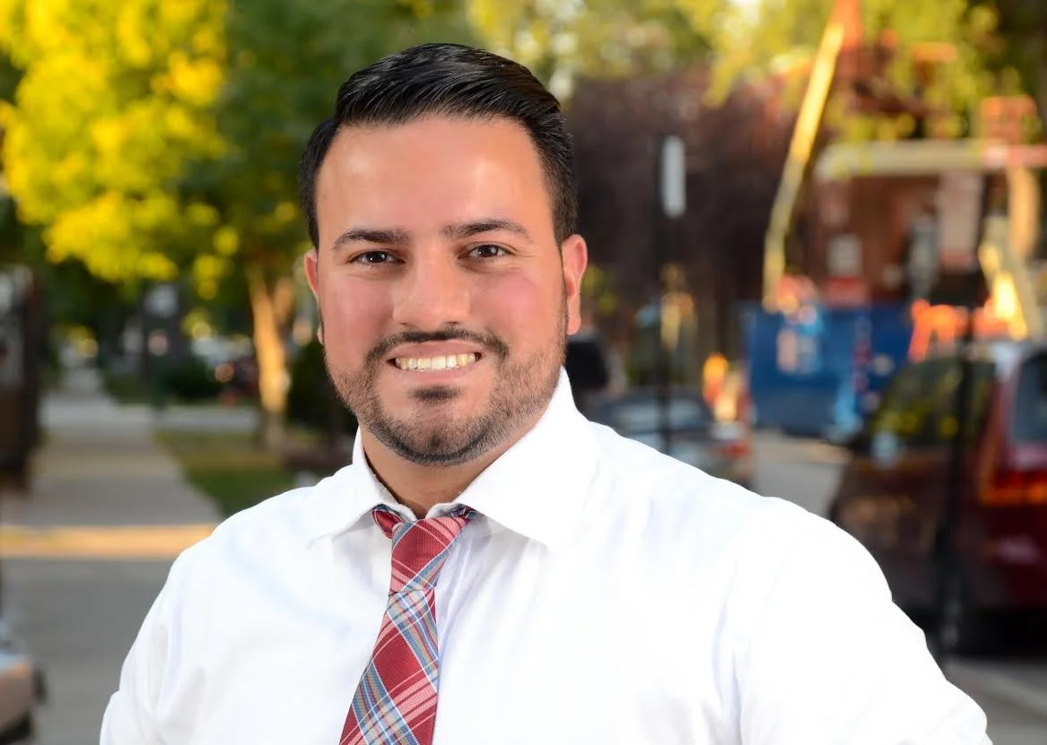 Portrait of a man smiling outdoors with trees and parked cars in the background.