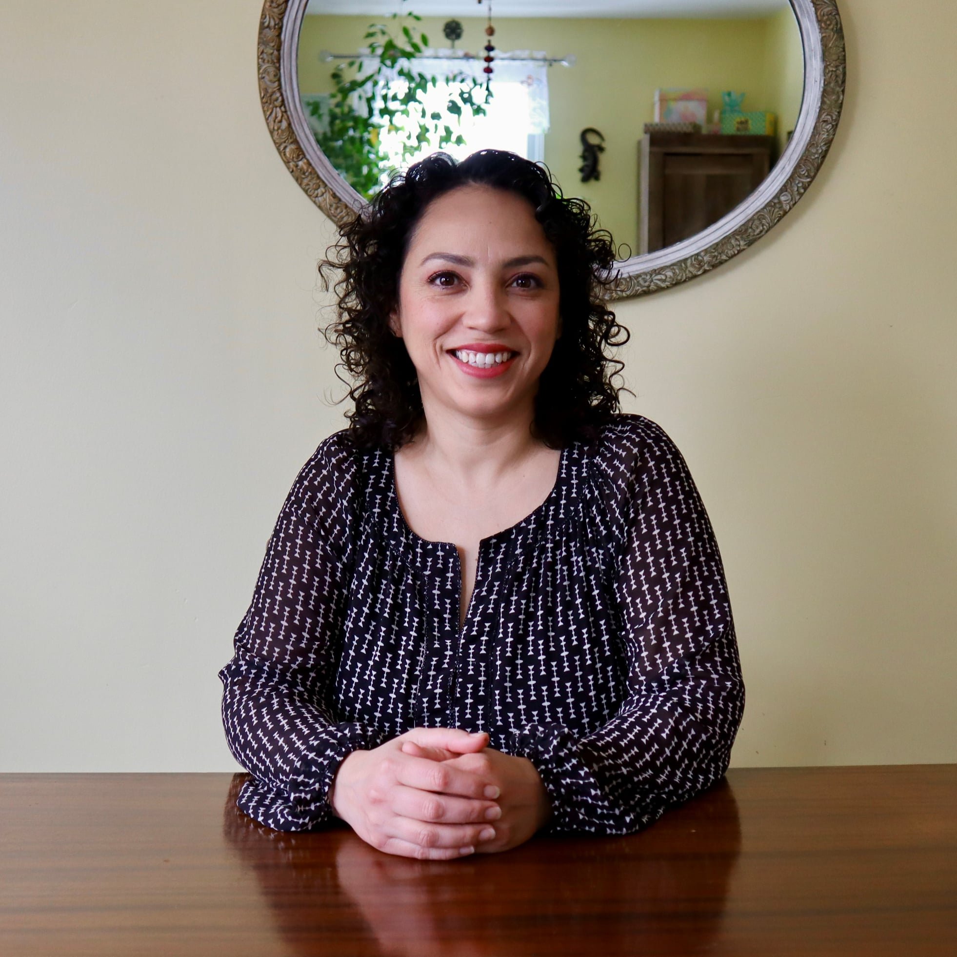 A woman with curly dark hair smiling, sitting at a wooden table with her hands clasped in front of her, in a room with a light yellow wall, behind her a large mirror with an ornate frame, some houseplants, and a wooden cabinet.